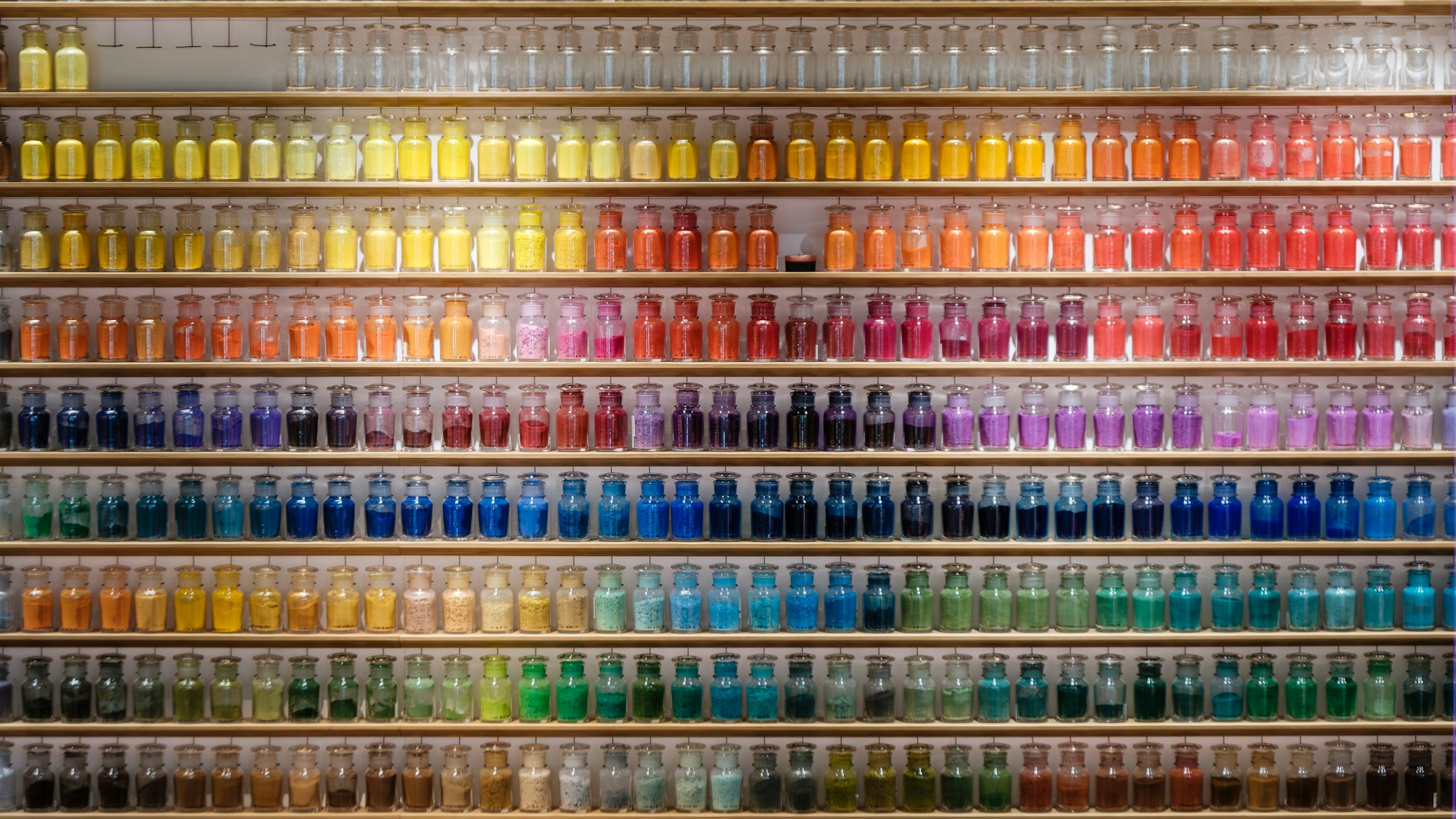 Scaffali di vetro con file ordinate di piccoli barattoli, ognuno contenente pigmenti colorati, creando un arcobaleno visivo che spazia dai toni gialli a quelli blu.