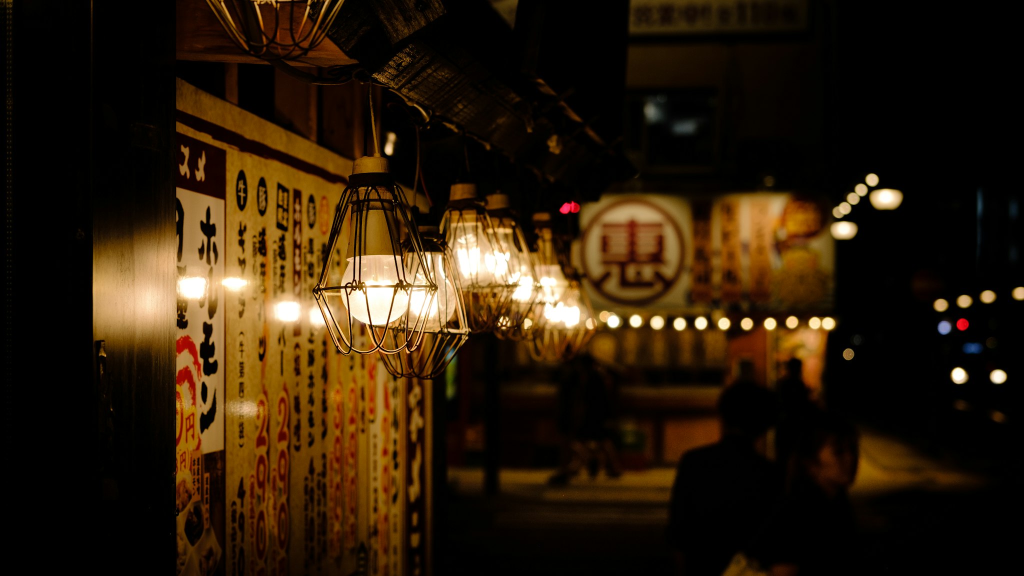 A dimly lit street scene in Japan features ornate lanterns glowing warmly, illuminating traditional signage adorned with intricate Japanese calligraphy, creating an inviting and atmospheric nighttime setting.
