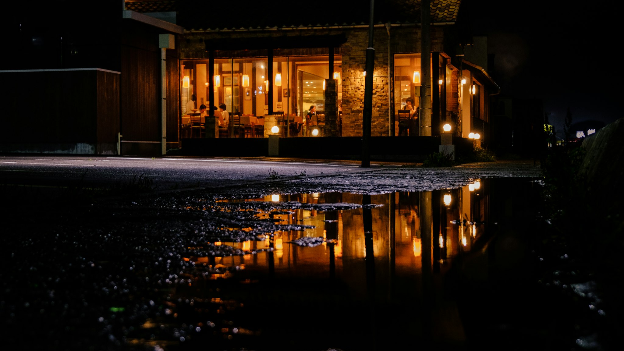 A cozy, warmly lit restaurant is seen at night across a wet street, with reflections glistening in puddles on the asphalt.