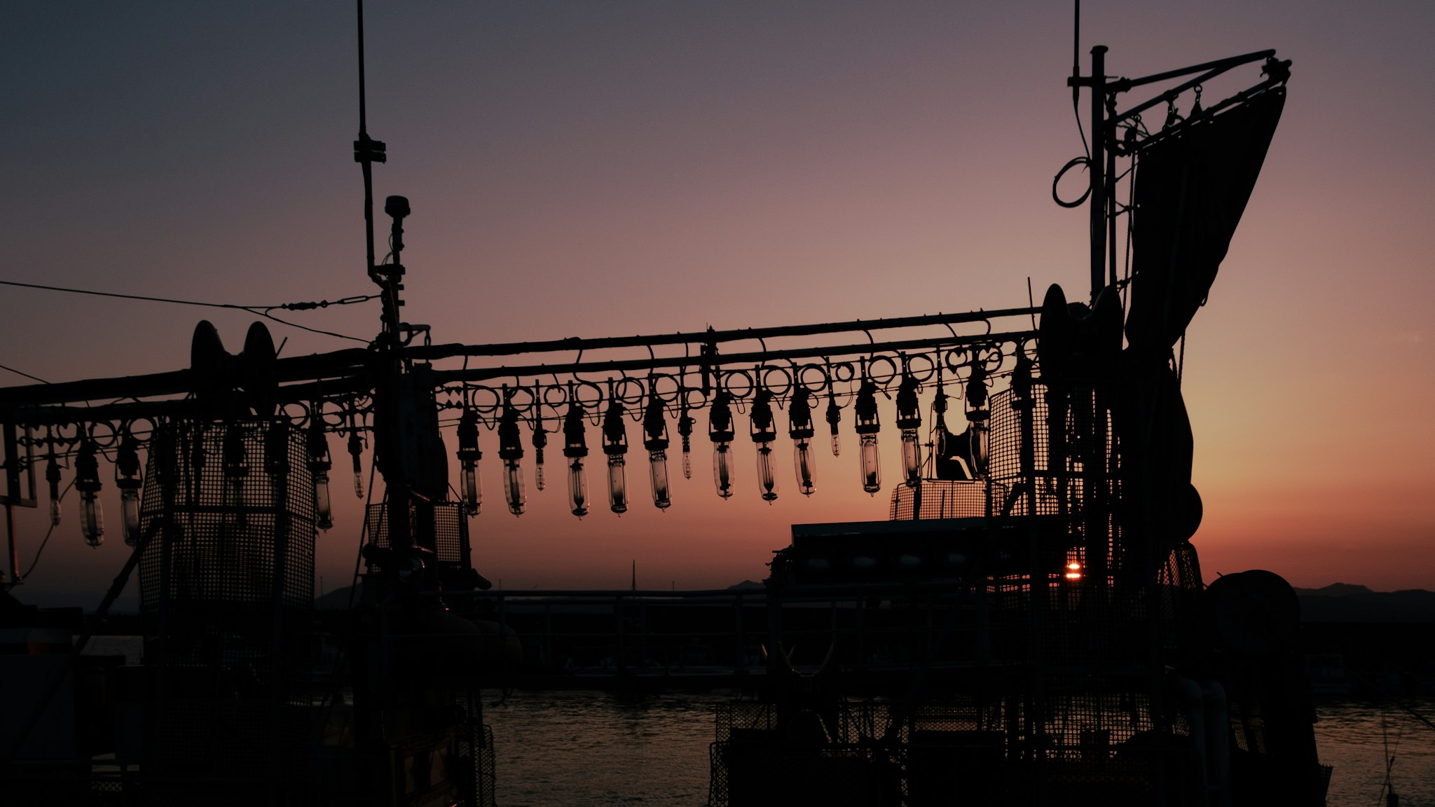 Silhouetted against a vibrant, pink-hued sunset, a fishing boat's intricate rigging and equipment are prominently featured, capturing a serene maritime scene at twilight.