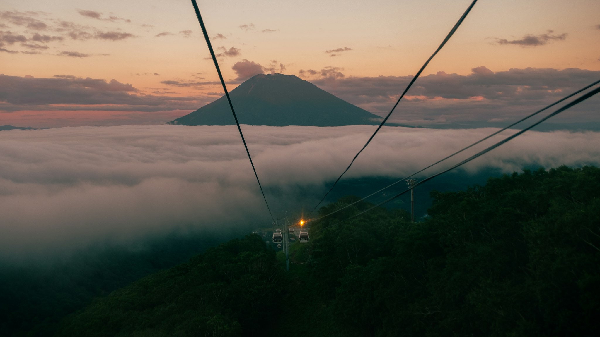 A cable car traverses over lush green mountains shrouded in mist, with a silhouetted volcano in the distance under a vibrant sunset sky.