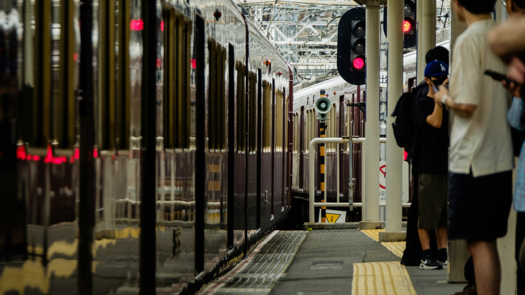 A bustling train station platform with passengers waiting beside a sleek, stationary train, as overhead lights and a digital signal board provide guidance.