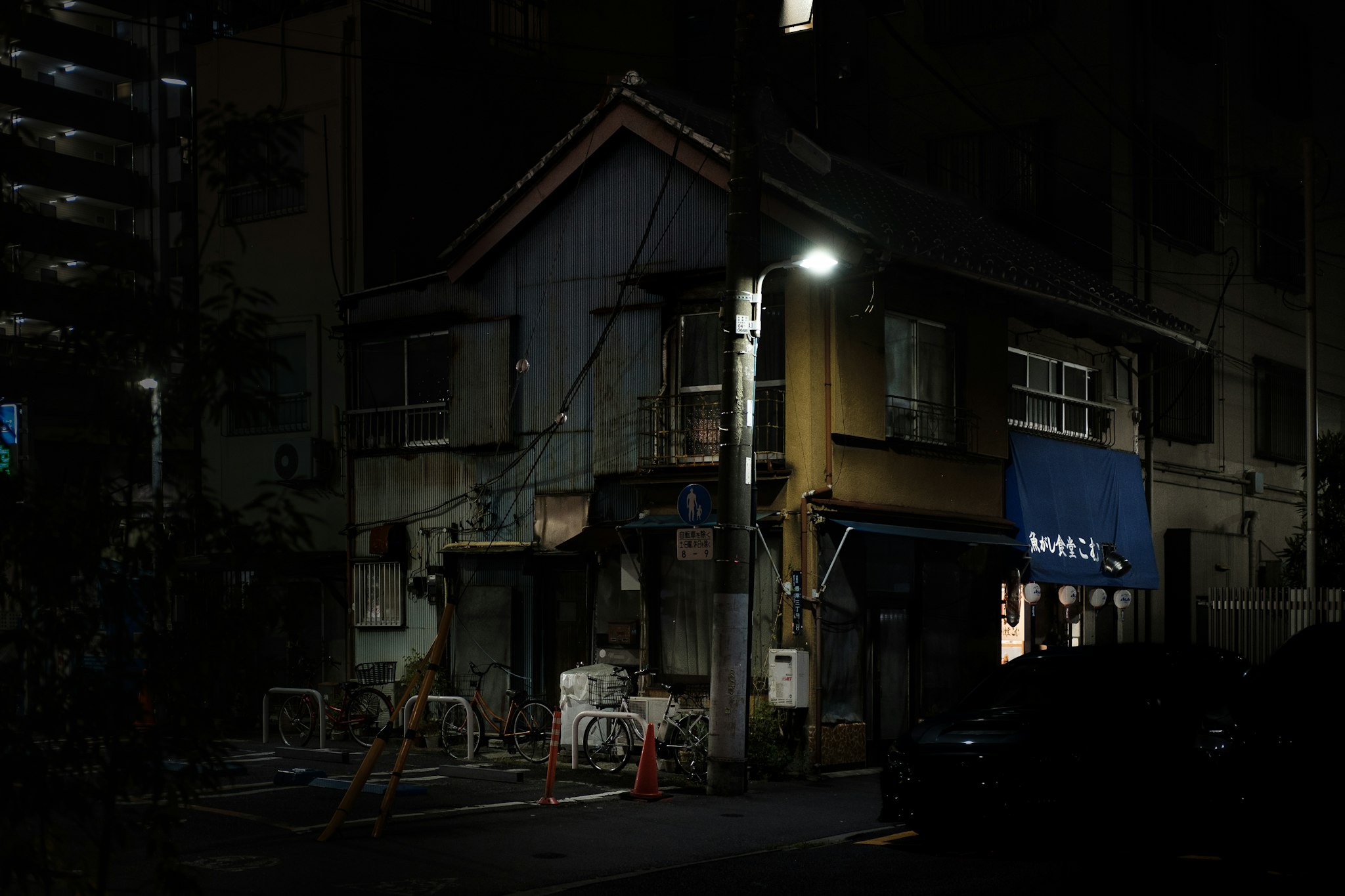 In una strada buia illuminata da un lampione, si trovano edifici tradizionali dall'aspetto rustico con insegne di stoffa blu, creando un'atmosfera tranquilla e suggestiva di un quartiere giapponese notturno.