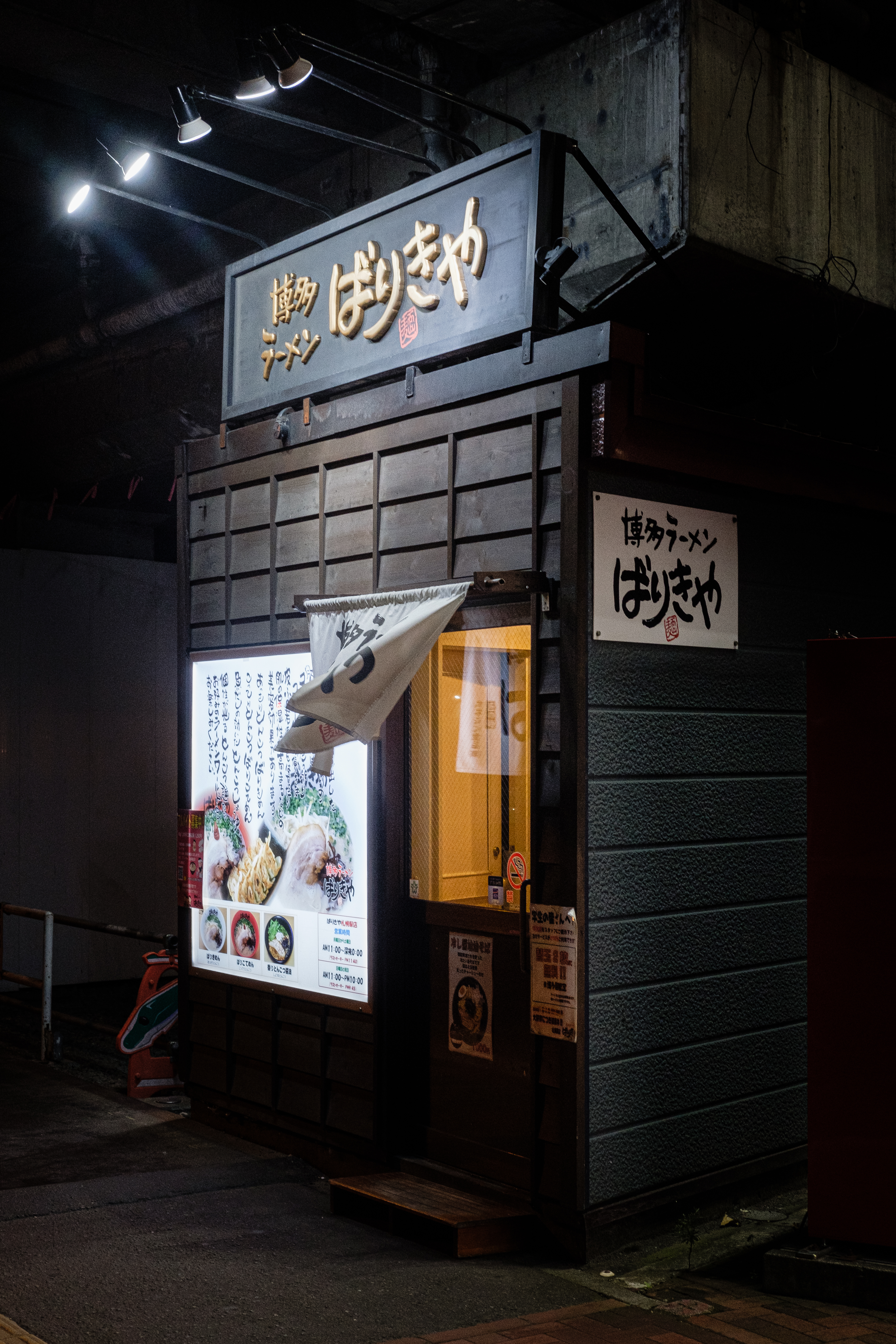 Esterno di un ristorante di ramen giapponese sotto un ponte, illuminato da luci soffuse, con insegne in giapponese e un menu esposto accanto alla porta d'ingresso.