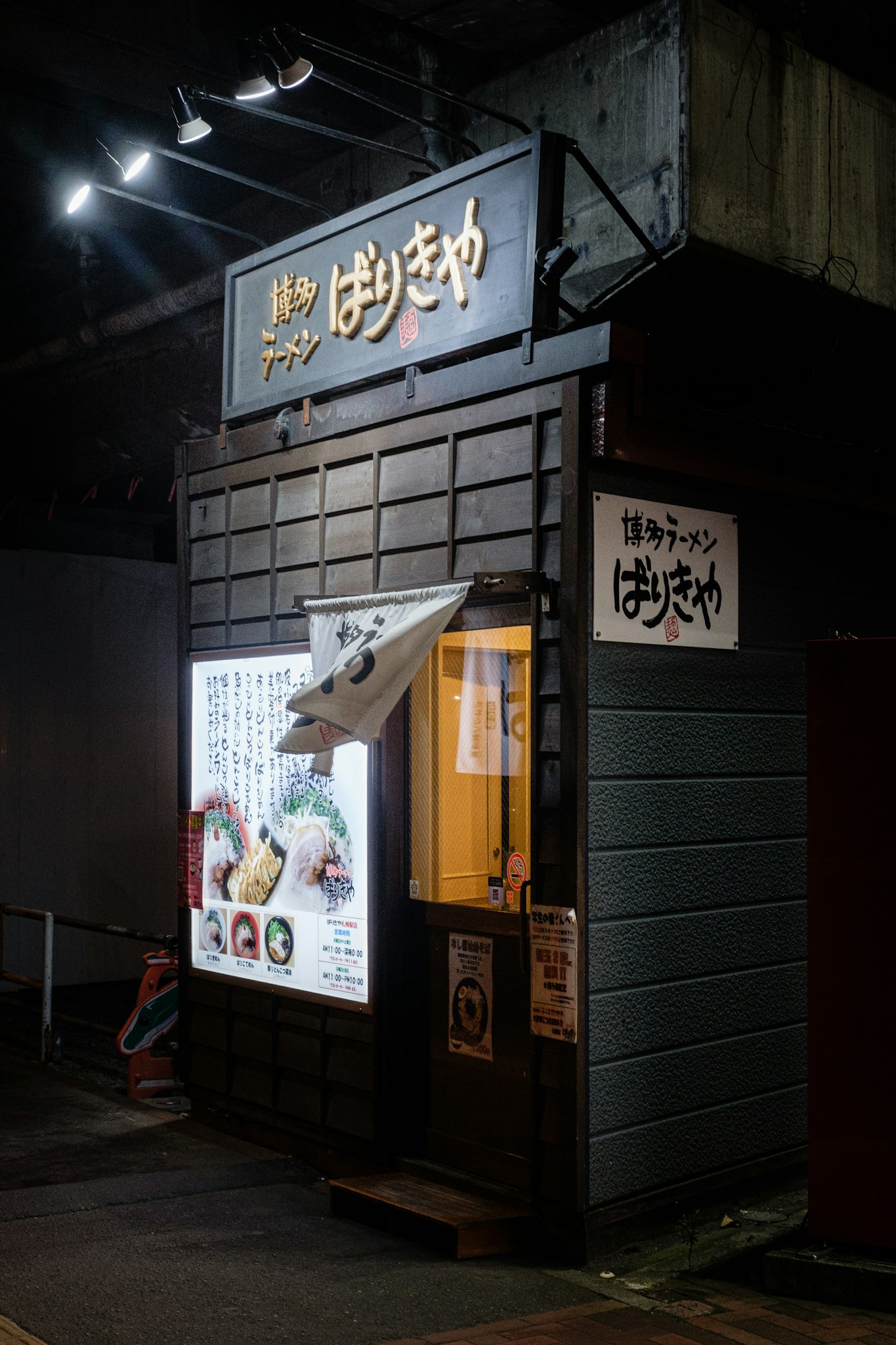A small, inviting ramen shop on a dimly lit street corner features a wooden facade with a sign in Japanese, a white fabric banner at the entrance, and illuminated pictures of menu items by the door, enhancing its authentic atmosphere.