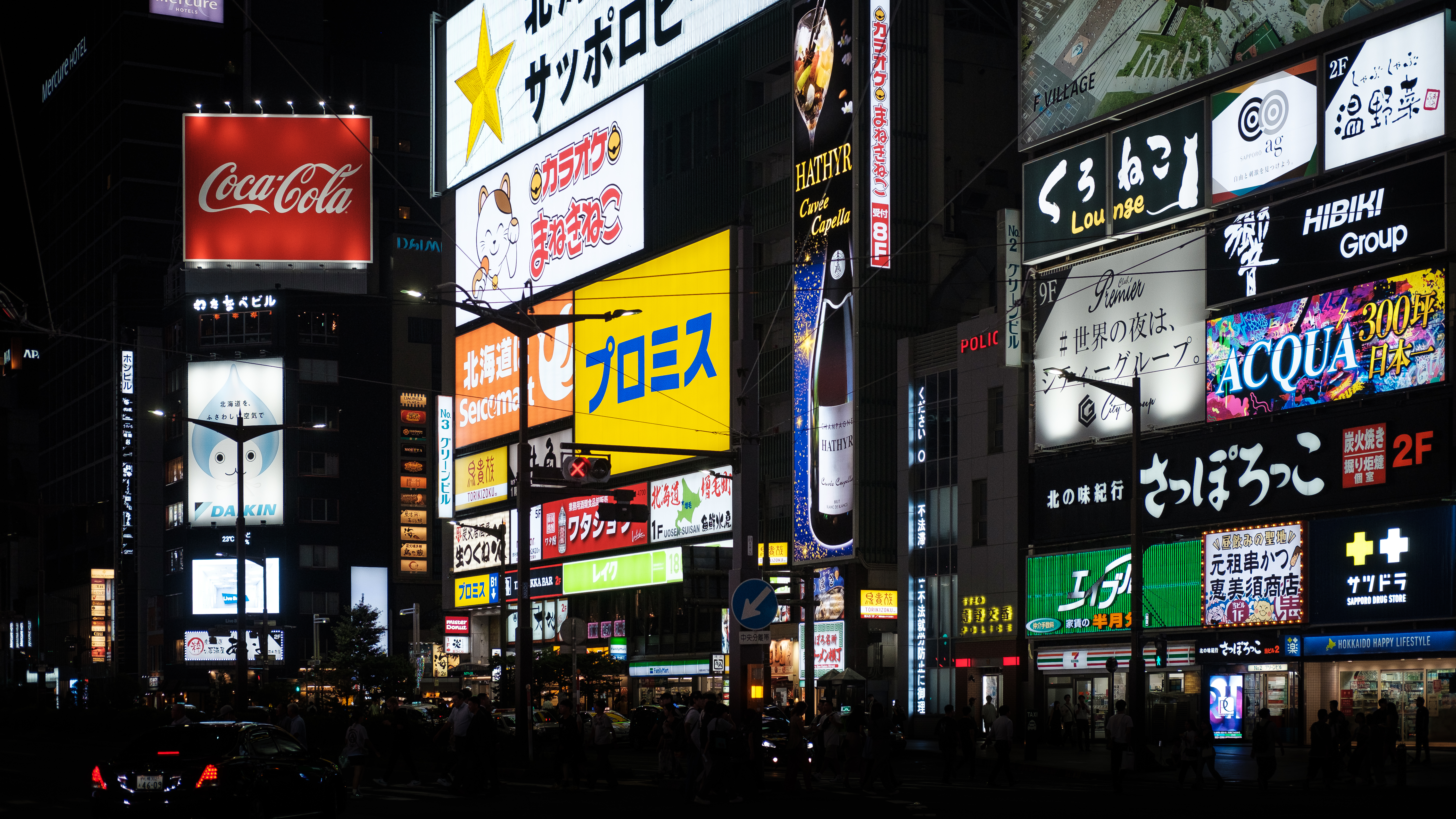Le luci al neon colorate dei cartelloni pubblicitari illuminano vividamente una strada affollata nel distretto commerciale di una grande città, con persone che camminano e auto che passano.