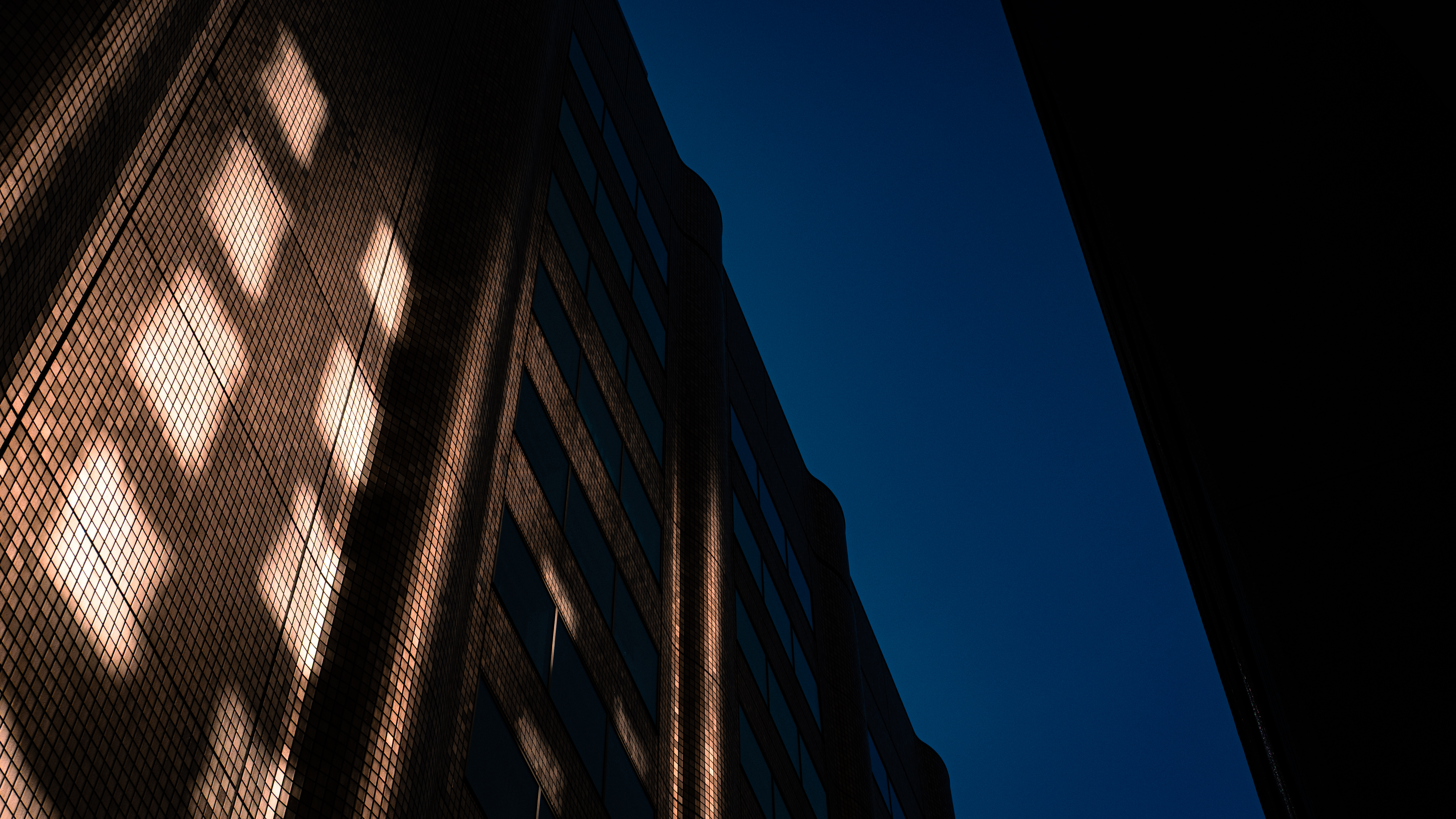Un imponente edificio in mattoni con finestre quadrate riflette la luce del sole al tramonto, creando giochi di luci e ombre sotto un cielo blu profondo in centro città.