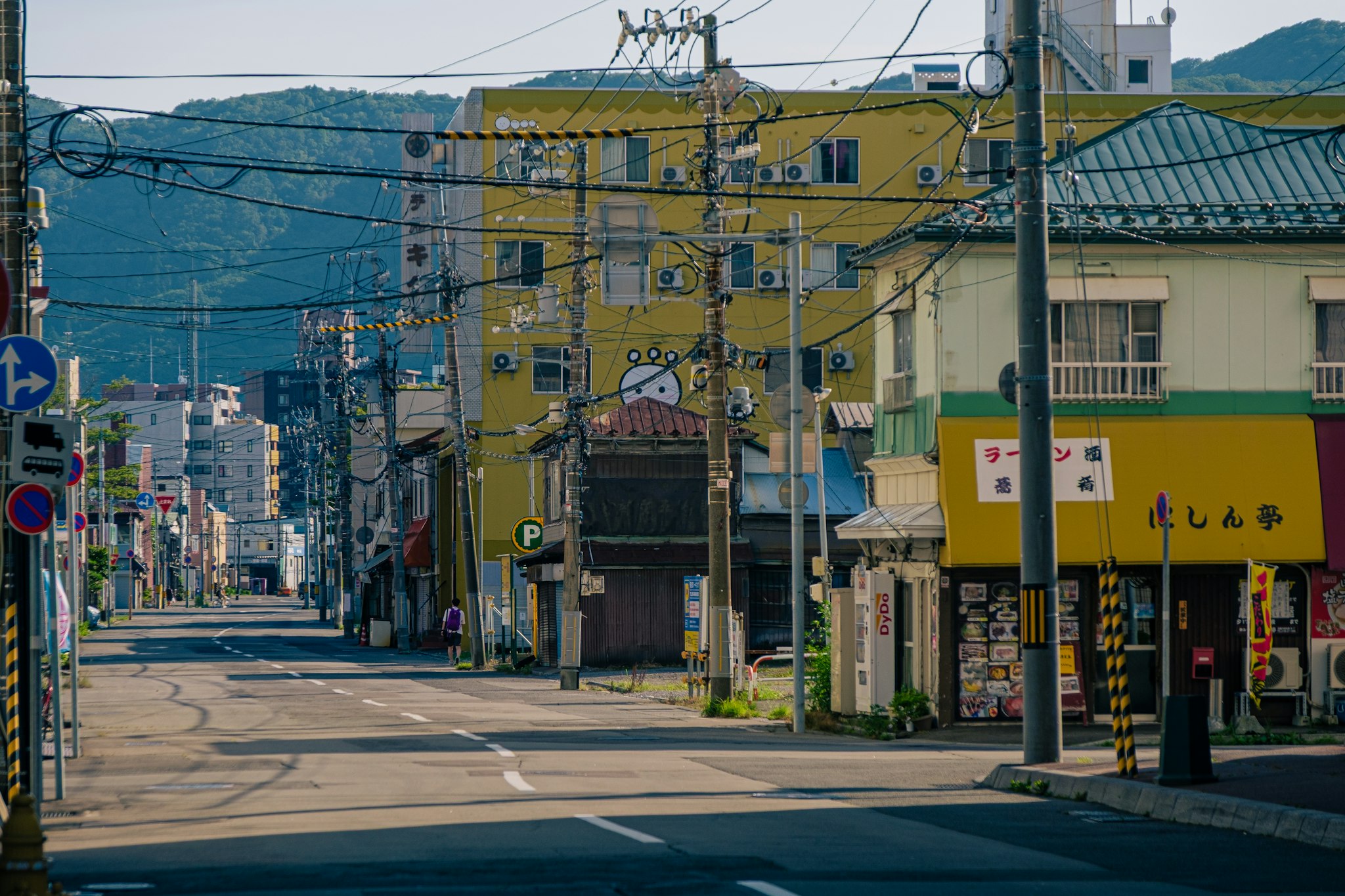 Una strada cittadina tranquilla in Giappone mostra edifici colorati e cartelli giapponesi, con cavi elettrici sospesi e montagne verdi sullo sfondo.