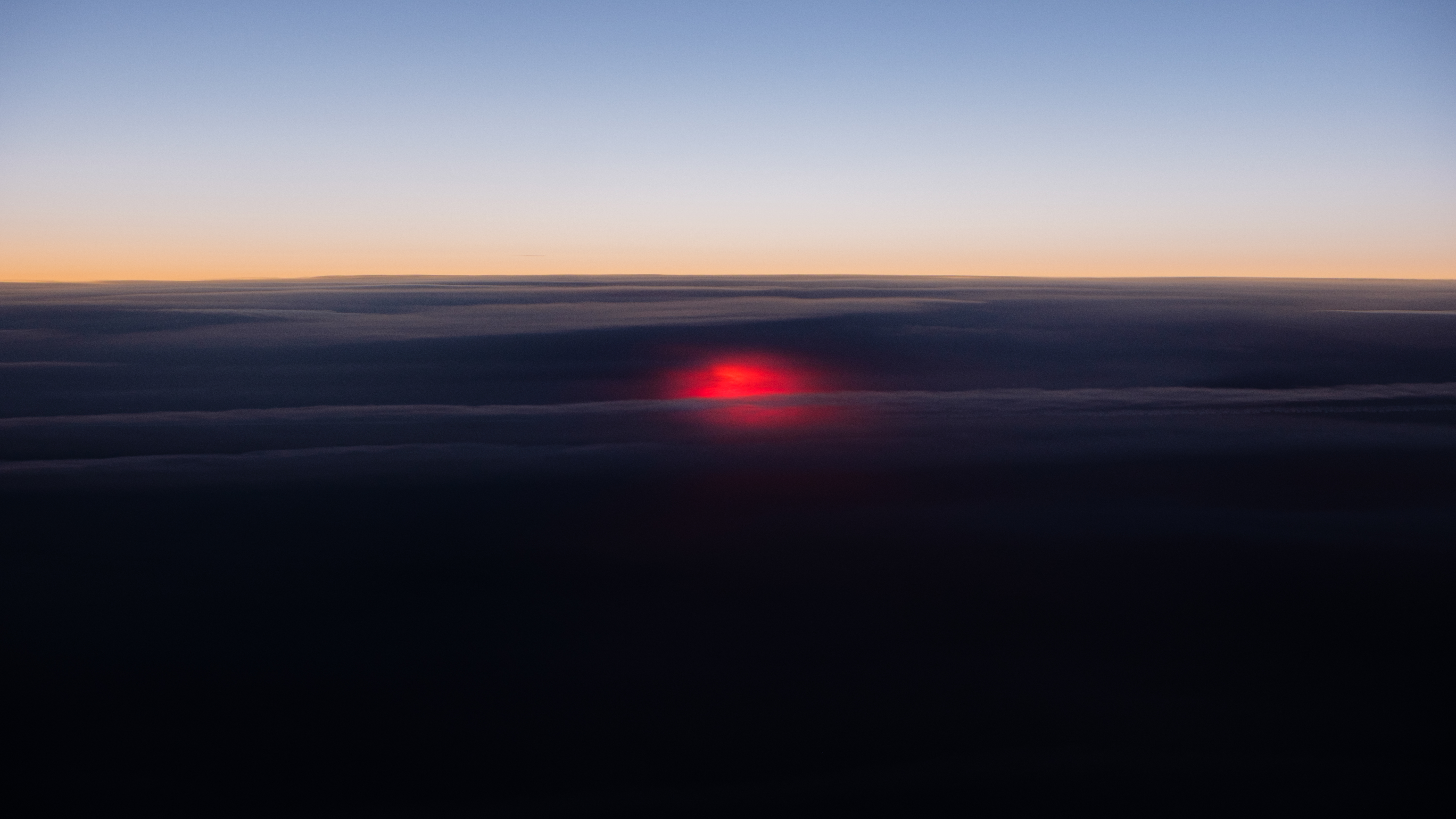 Il sole che tramonte visto da sopra le nuvole, con un bagliore rosso che illumina le nuvole al centro, creando un contrasto tra il cielo blu e le tonalità scure delle nuvole sottostanti.