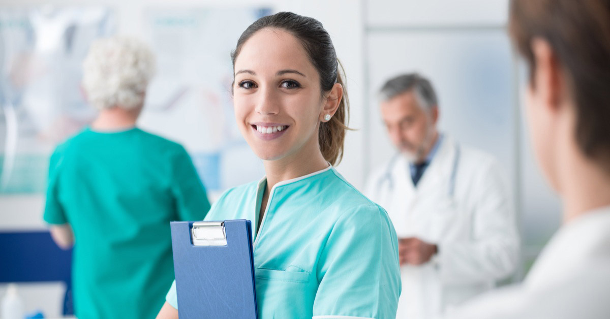 A young nurse smiles while holding a clipboard to her chest within a hospital