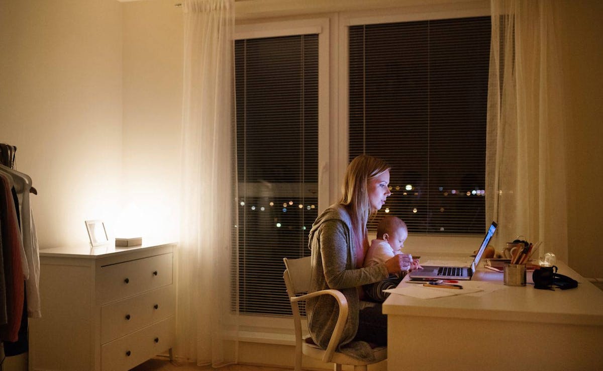 A young mother sits at a desk in front of her laptop at night. Her baby is seated in her lap.