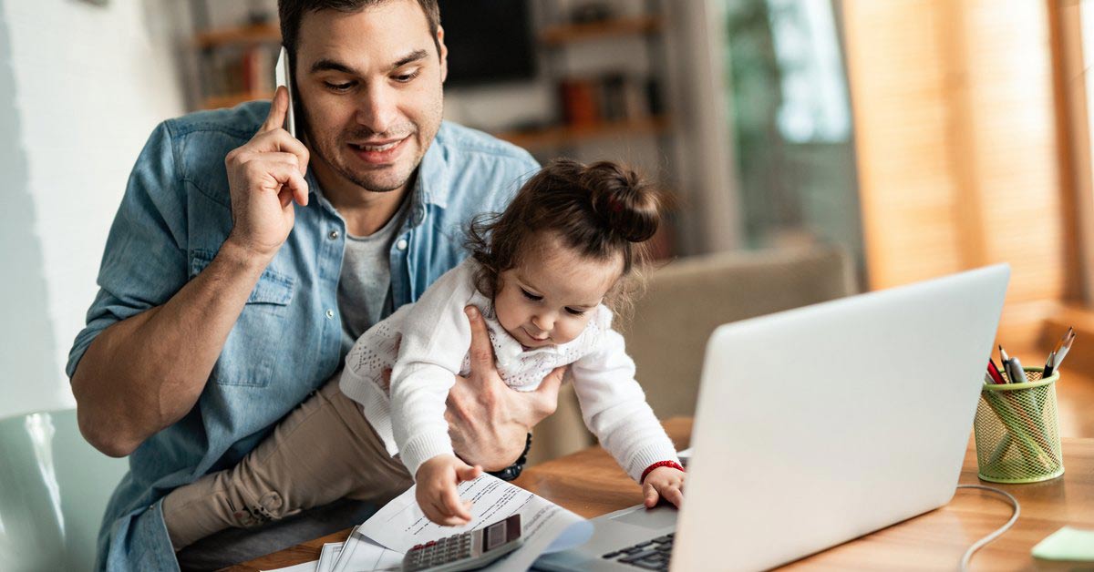 A father holds his child in his lap while he takes a phone call. His laptop is open on a desk in front of him.