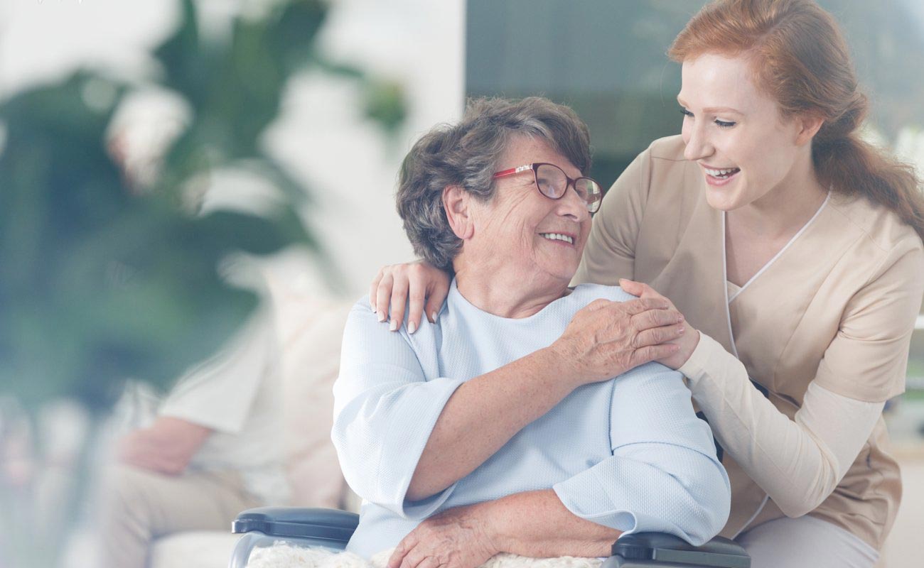 An elderly woman reaches over her shoulder, smiling, to laugh with a red-haired female health worker behind her