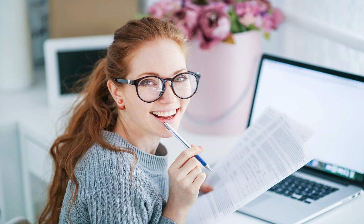 A young red-headed woman wearing glasses is seated in front of her open laptop and smiles into the camera