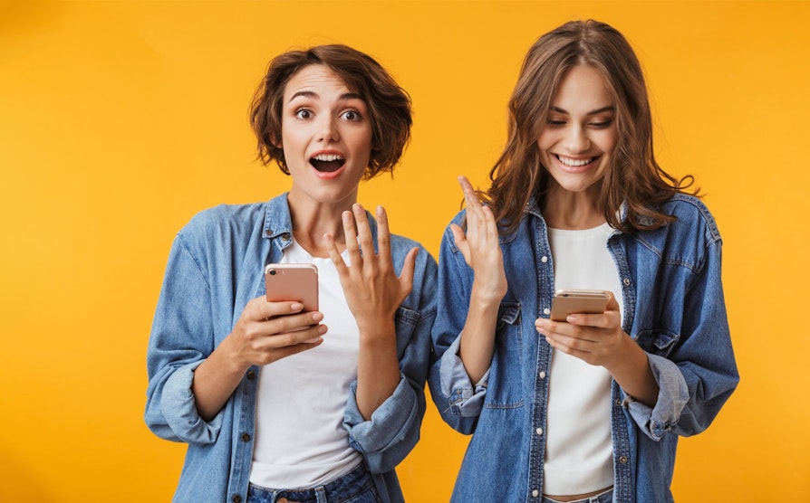 To glade jenter med mobiltelefoner; en av de ser i kamera med et overrasket smil.