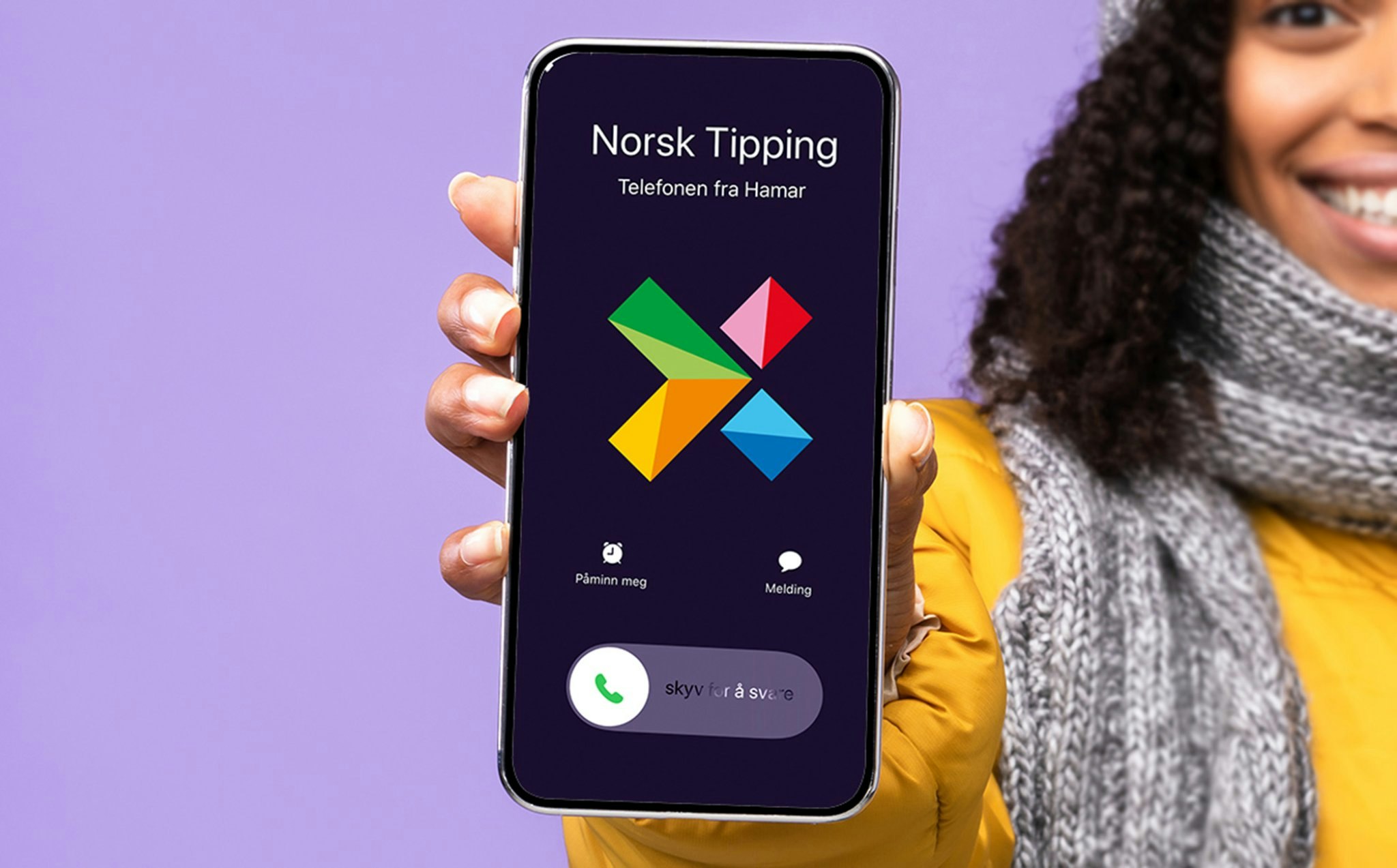Kvinne med gul boblejakke og grått skjerf smiler og holder opp en mobiltelefon som viser et anrop fra Norsk Tipping mot lilla bakgrunn.