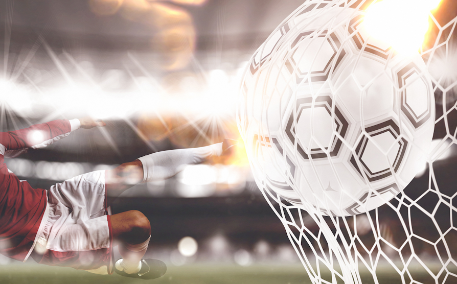 En fotballspiller som skyter ballen i mål. Fotballspilleren i rød drakt og hvit shorts.