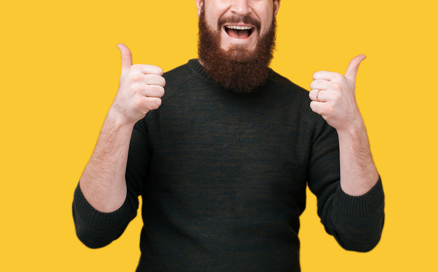 Utnsitt av halve hodet og overkroppen til en mann med brunt skjegg og grå genser som smiler og holder to tomler opp mot gul bakgrunn.