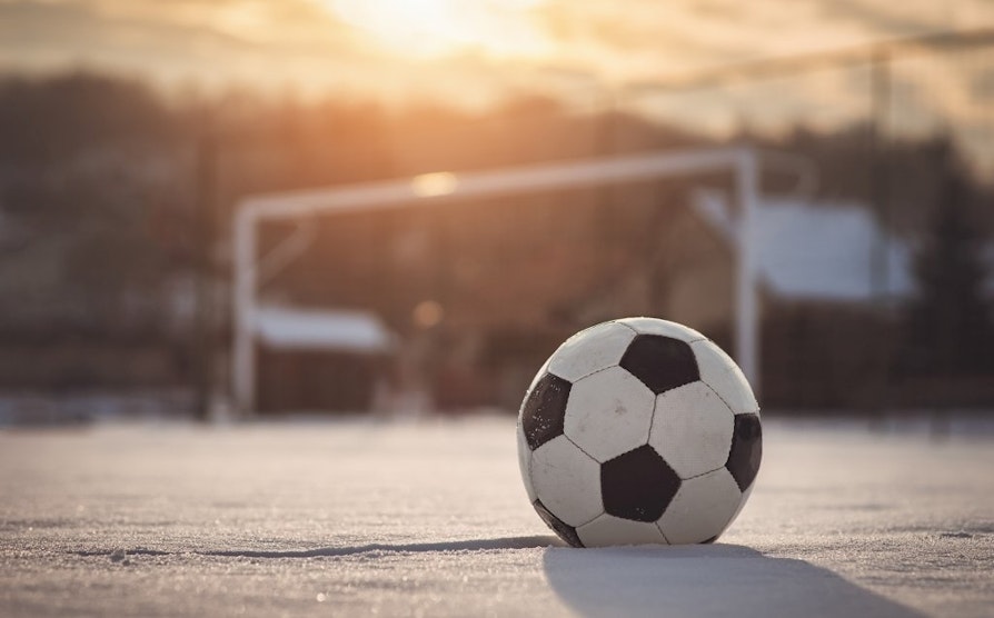 Illustrasjonsfoto av fotball som ligger ute på snødekt bane; et mål står ufokusert i bakgrunnen