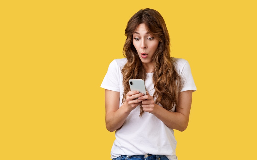 Illustrasjonsfoto av kvinne på gul bakgrunn som stirrer overrasket på mobiltelefonen.