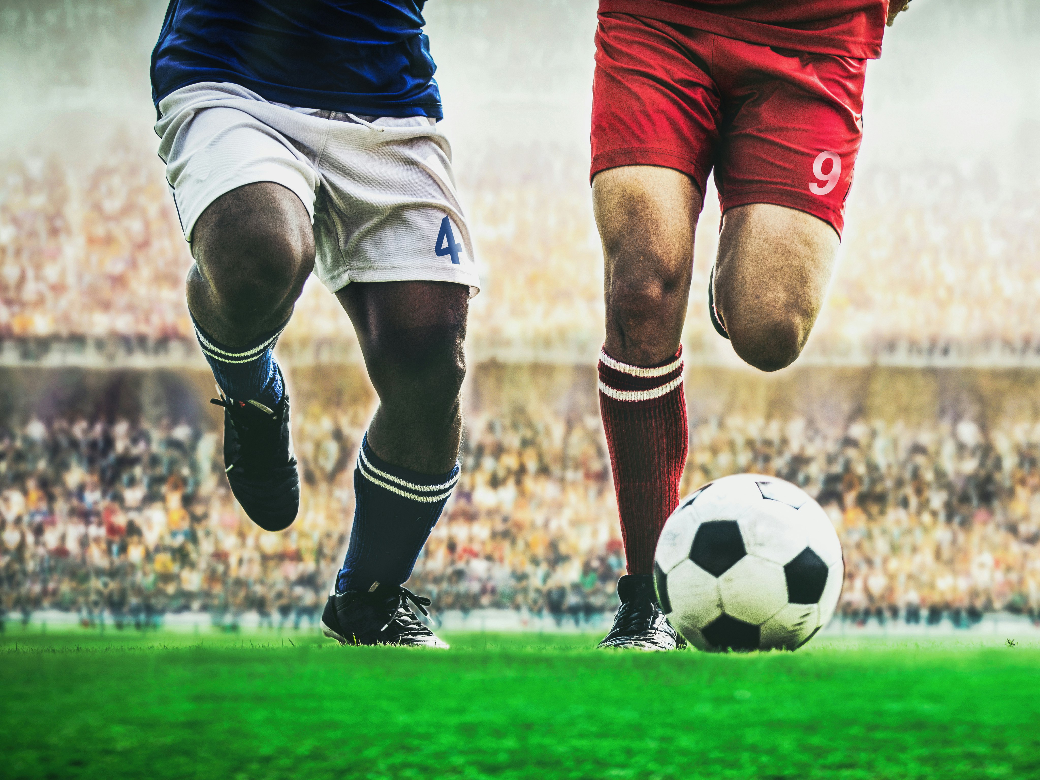 Illustrasjonsfoto av beina til to fotballspillere som jager en fotball på gressbane: den ene har blå og hvit drakt, den andre har rød
