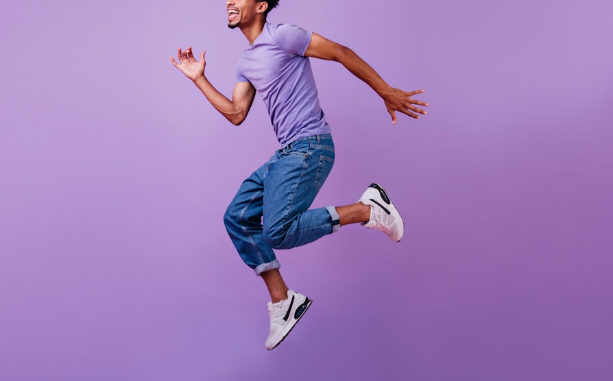Mann i lilla t-skjorte, jeans og joggesko som hopper med lilla bakgrunn.