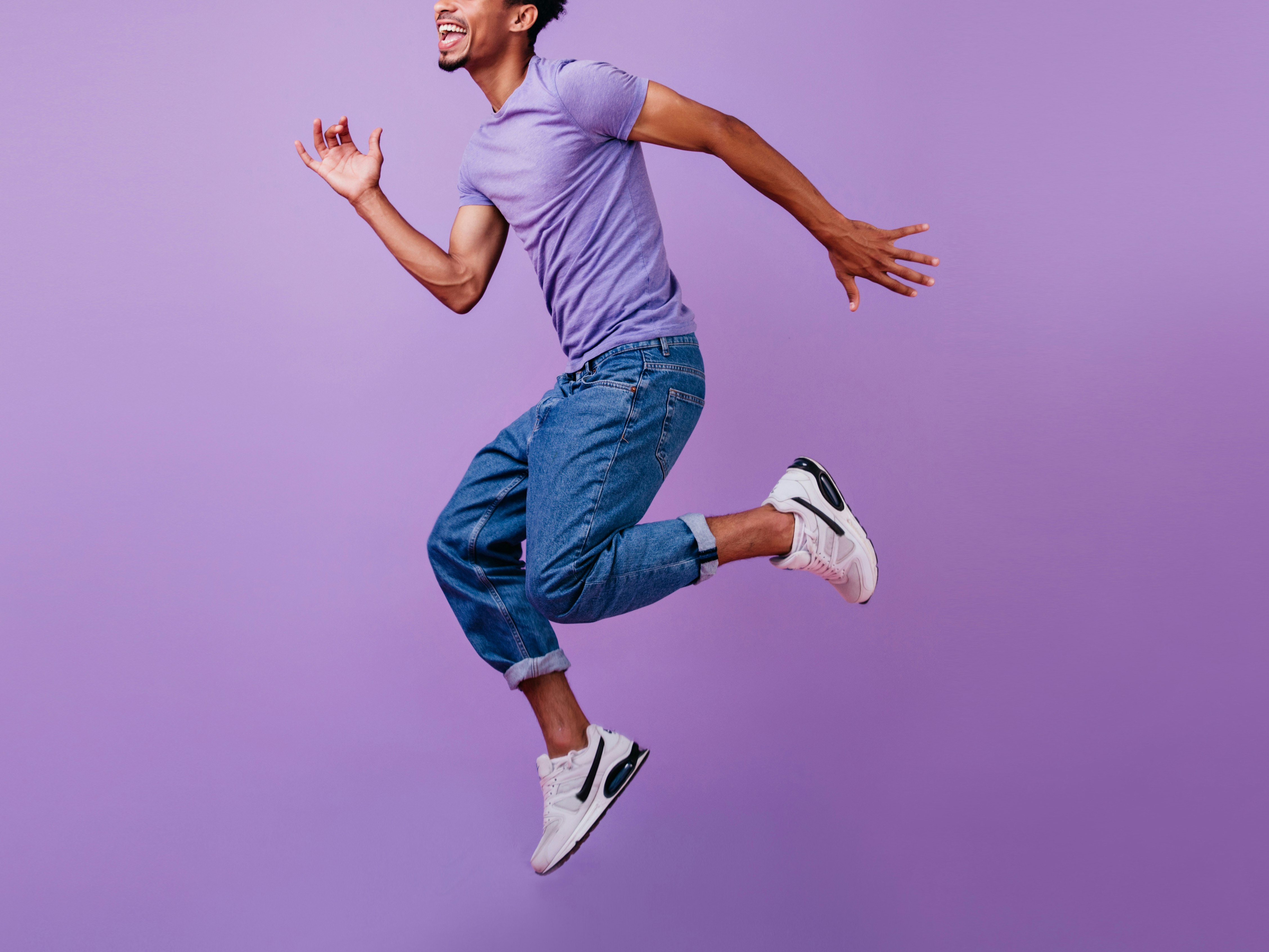 Mann i lilla t-skjorte, jeans og joggesko som hopper med lilla bakgrunn.