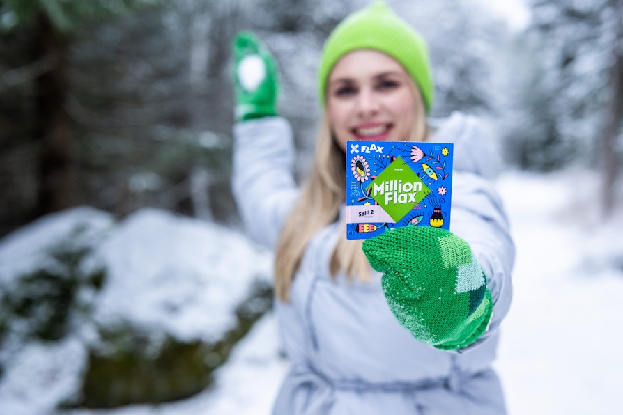 Smilende blond kvinne i vinterskog ikledd  blå boblejakke og grønne votter og lue holder et MillionFlax-lodd i den ene hånden og en snøball i den andre.