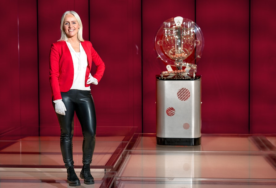 Bilde av kvinne, Ingrid Roterud Mathisen i Norsk Tipping. Står i tv-studioet der Lotto-trekningen spilles inn. I bakgrunnen sees ny trekningsmaskin for Lotto. Rød bakgrunn.