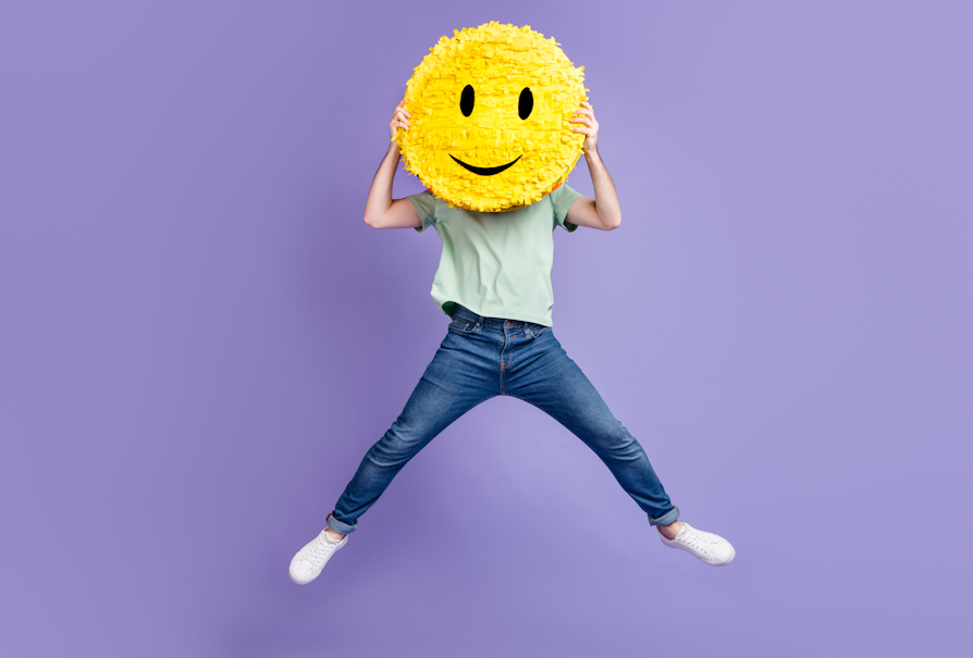 Mann i blå jeans, turkis t-skjorte og hvite joggesko holder en stort gult smile-face over ansiktet mens han hopper foran lilla bakgrunn
