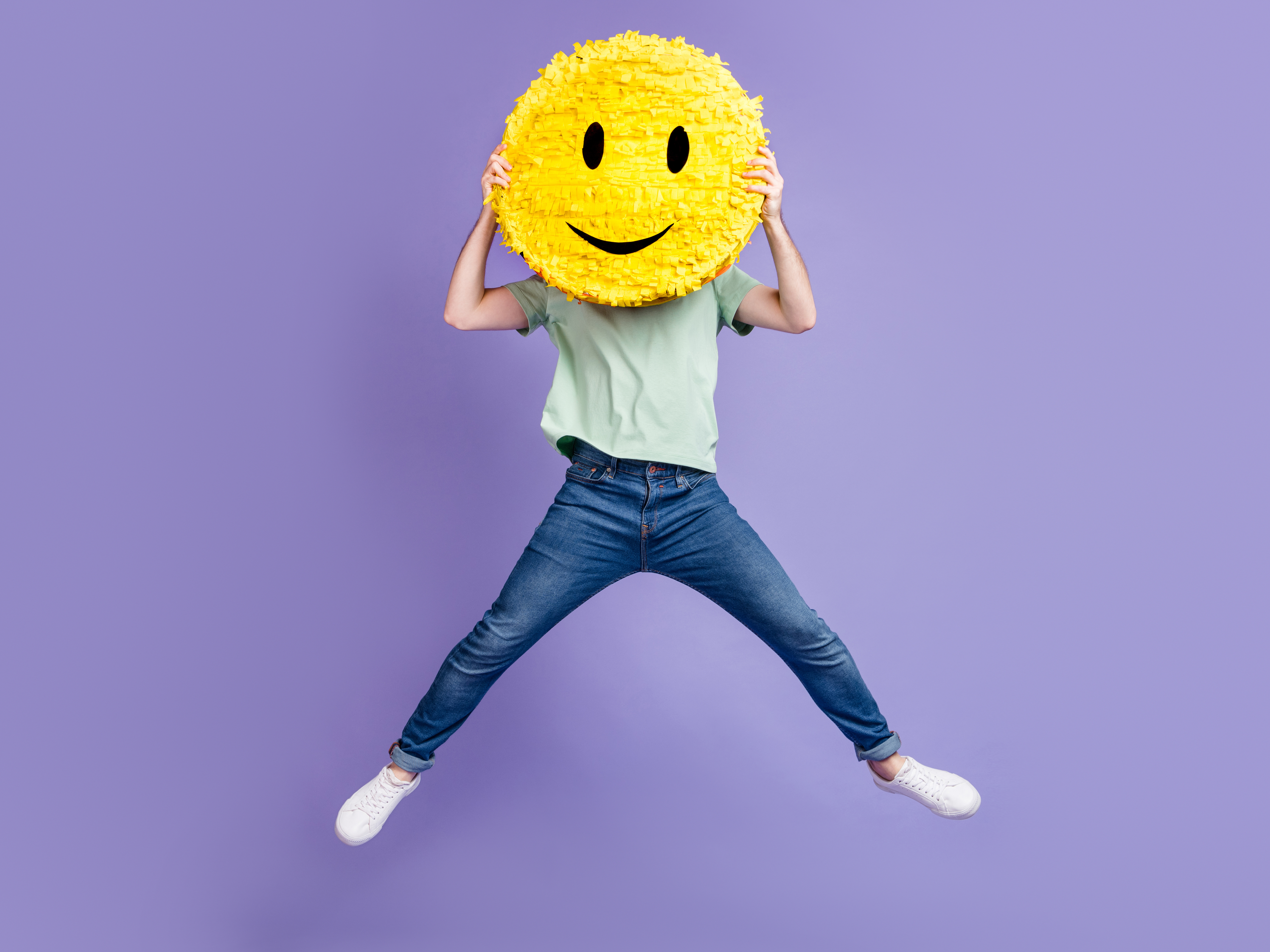 Mann i blå jeans, turkis t-skjorte og hvite joggesko holder en stort gult smile-face over ansiktet mens han hopper foran lilla bakgrunn