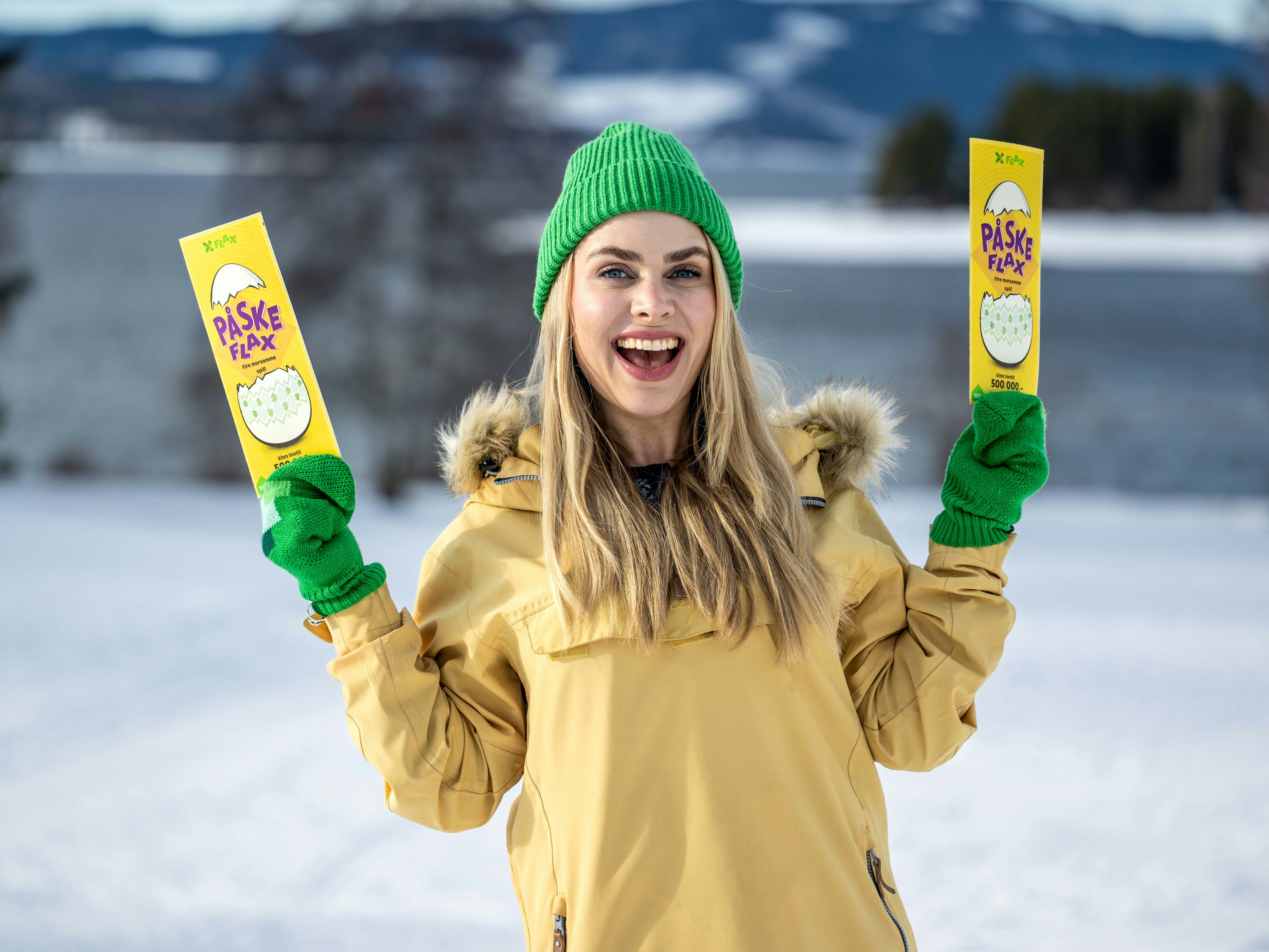Smilende kvinne i gul anorakk, grønn lue og grønne votter holder et PåskeFlax-lodd i hver hånd i vinterlandskap.