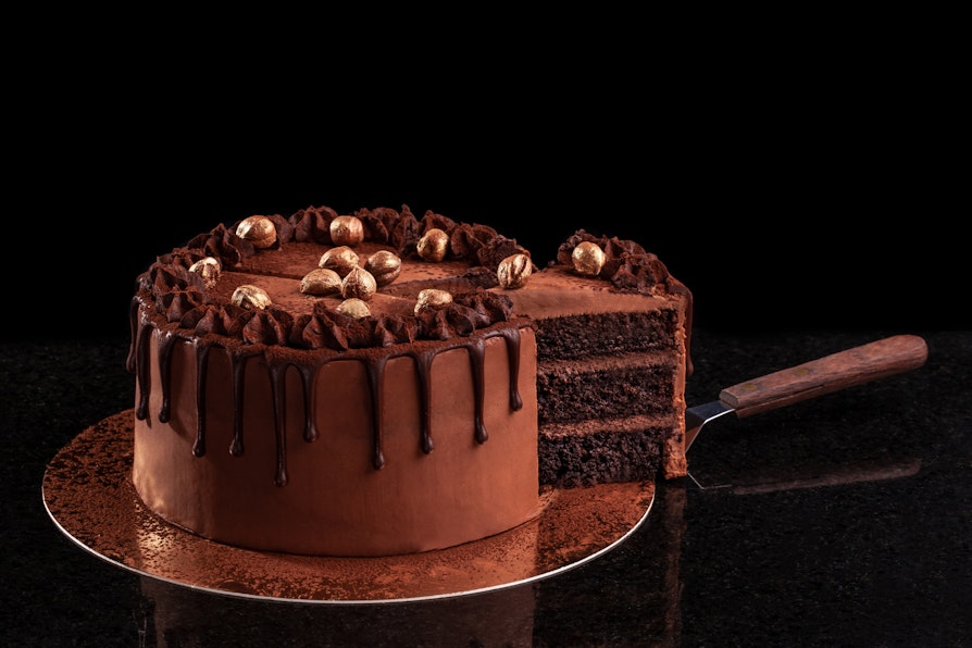 Bilde av en sjokoladekake på gullfat med sort bakgrunn hvor det en kakespade har kuttet opp en bit