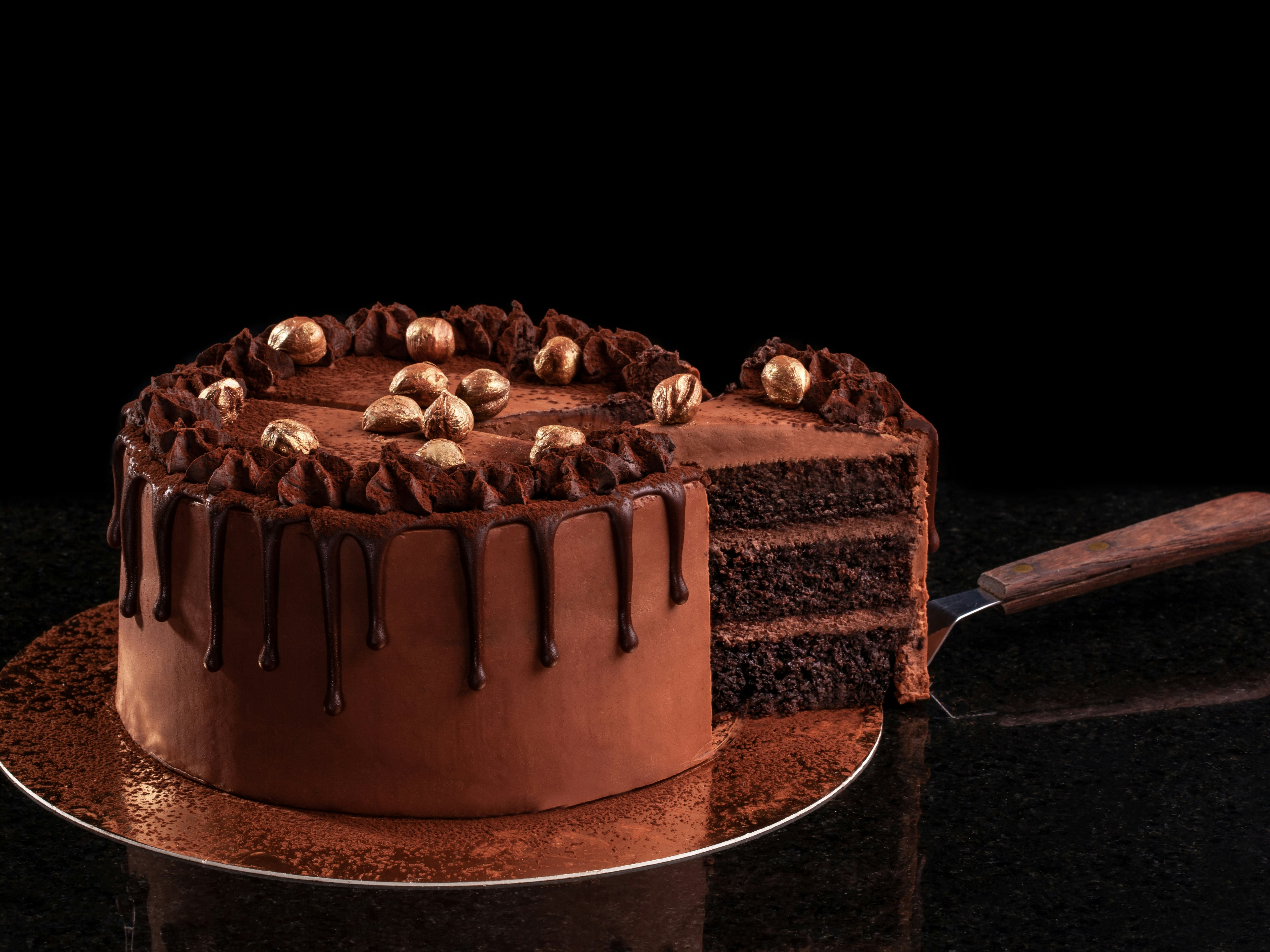 Bilde av en sjokoladekake på gullfat med sort bakgrunn hvor det en kakespade har kuttet opp en bit
