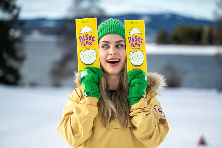Kvinne ikledd gul anorakk, grønne votter og grønn lue i vinterlige omgivelser, holder ett PåskeFlax-lodd i hver hånd ved siden av ansiktet