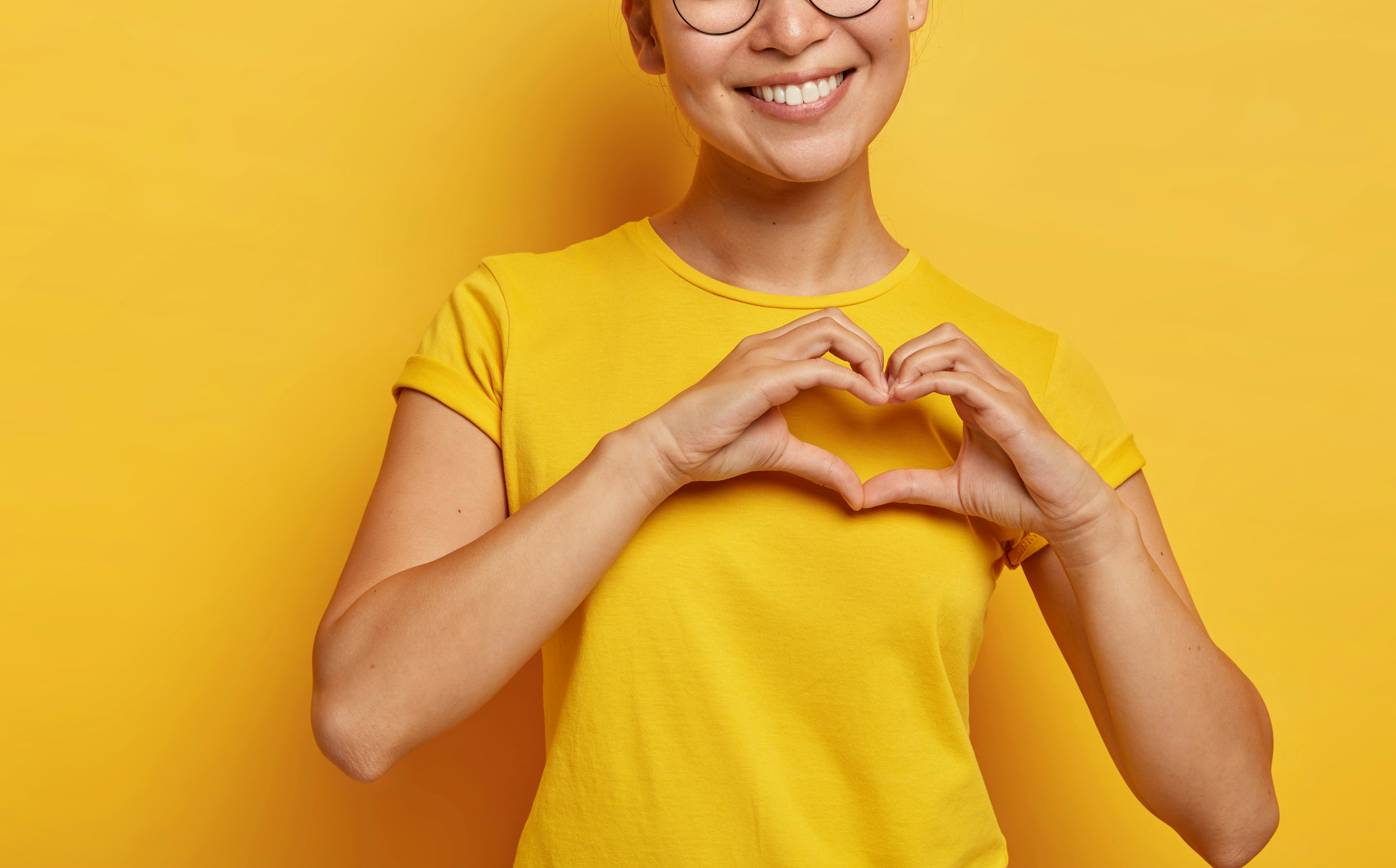 Smilende kvinne med briller og gul t-skjorte holder hendene sammen som et hjerte foran brystet mot en gul bakgrunn.