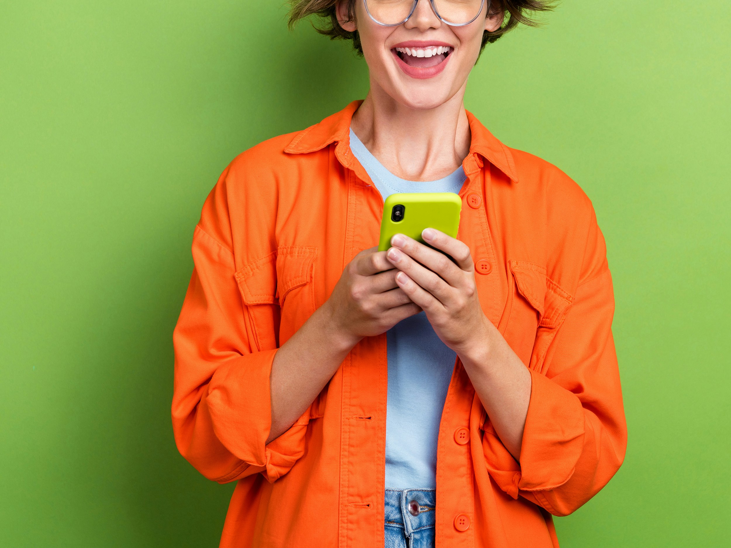 Illustrasjonsfoto (utsnitt) av smilende kvinne med mobiltelefon og oransje jakke på grønn bakgrunn