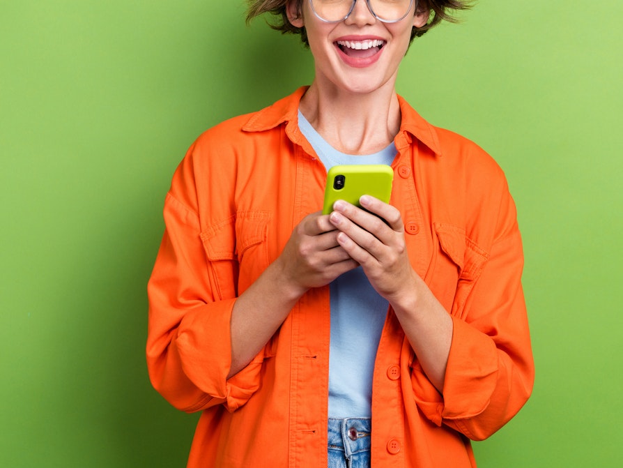 Illustrasjonsfoto (utsnitt) av smilende kvinne med mobiltelefon og oransje jakke på grønn bakgrunn