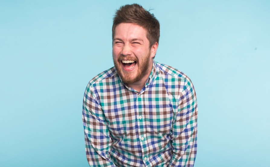 Mann med skjegg og rutete skjorte lener seg frem i latter foran lyseblå bakgrunn.