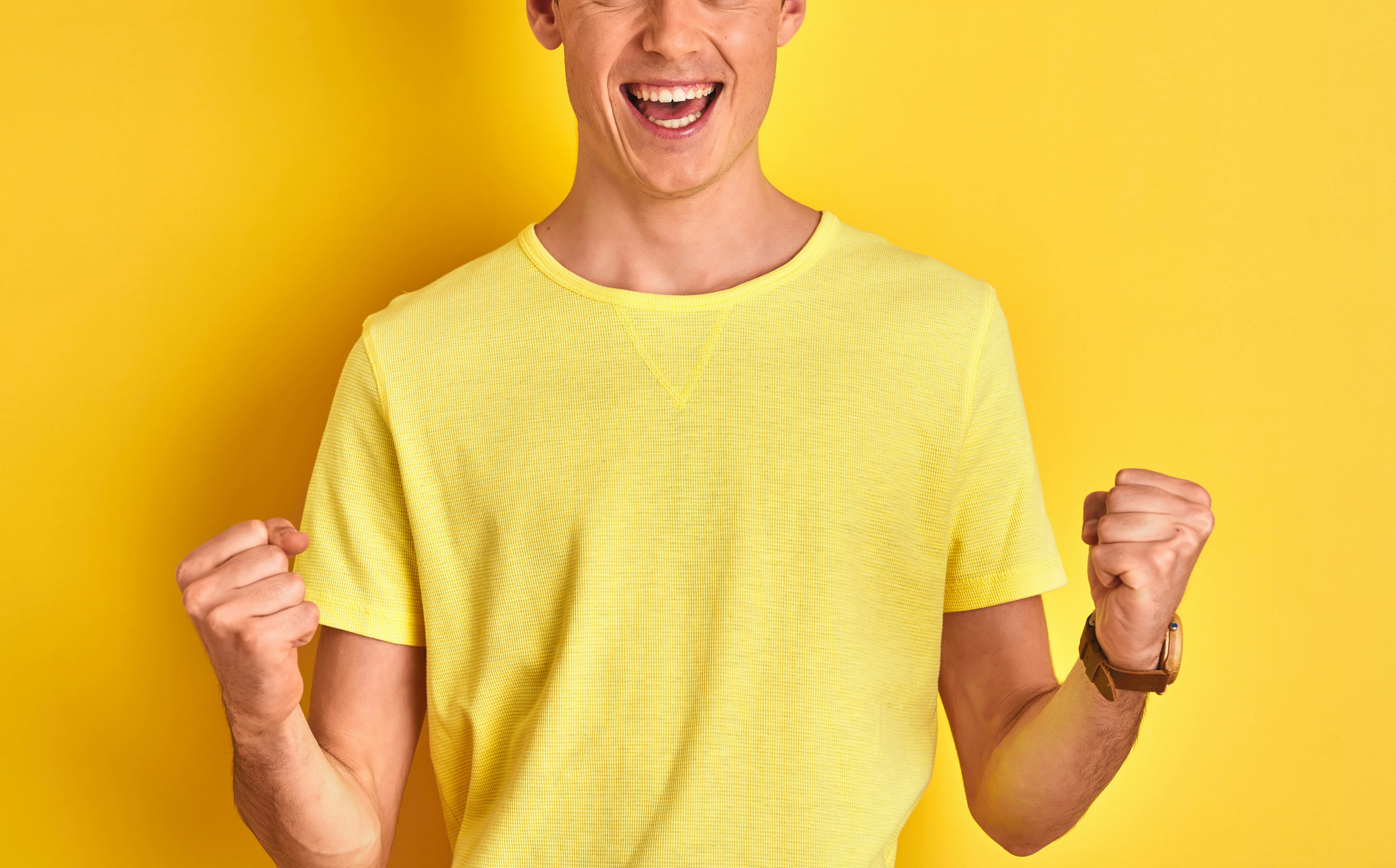 Mann i gul t-skjorte foran gul bakgrunn jubler og knytter hendene.