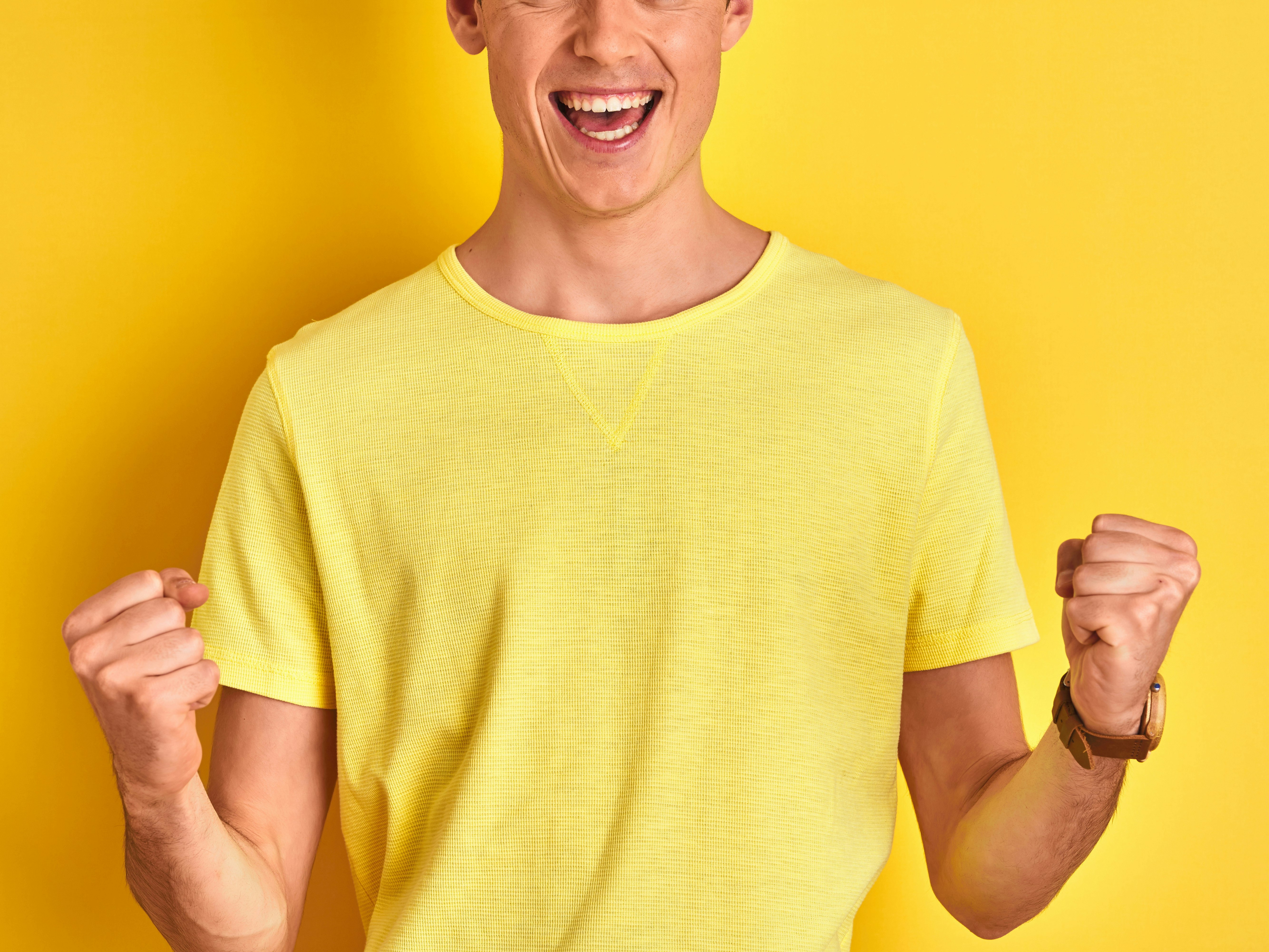 Mann i gul t-skjorte foran gul bakgrunn jubler og knytter hendene.