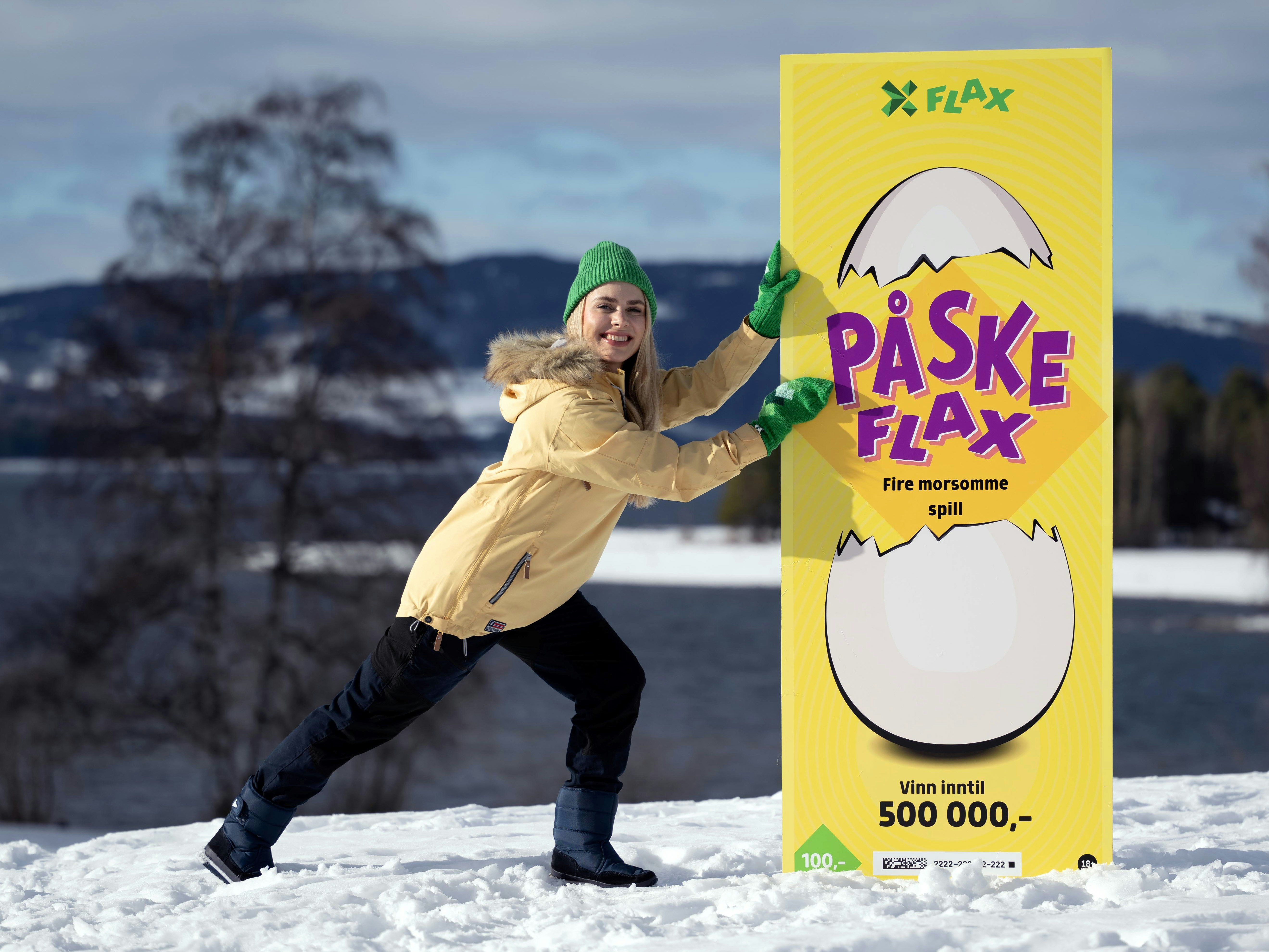 Kvinne i gul anorakk og grønn lue og votter støtter opp et overdimensjonert PåskeFlax-lodd i vinterlandskap.