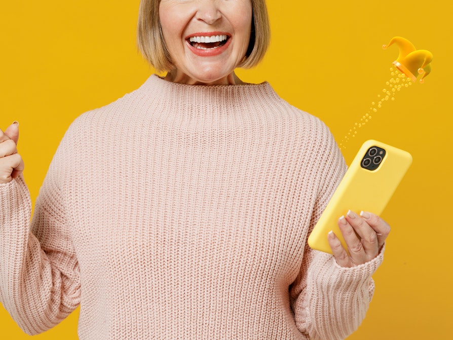 Illustrasjonsbilde av jublende kvinne på gul bakgrunn som holder en mobiltelefon, en Joker-lue flyter over