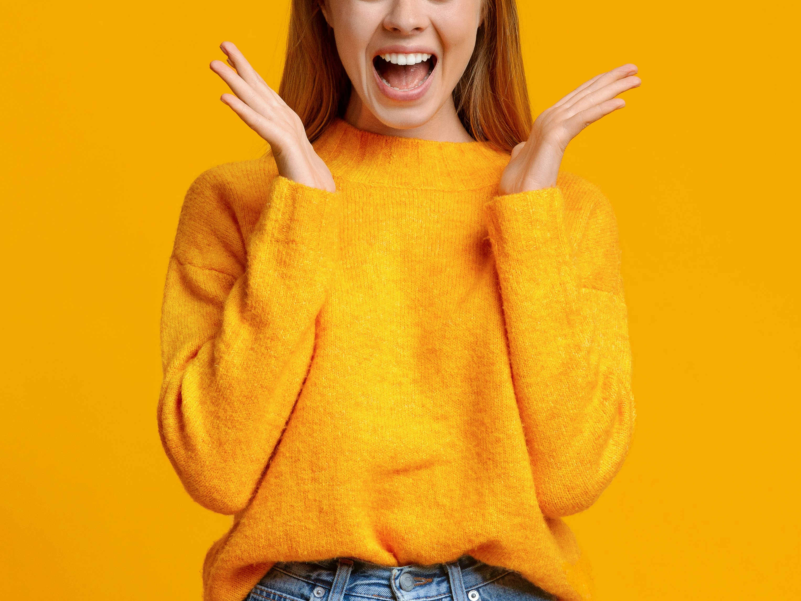 Blond kvinne i orange genser med overrasket uttrykk og åpen munn mot orange bakgrunn.
