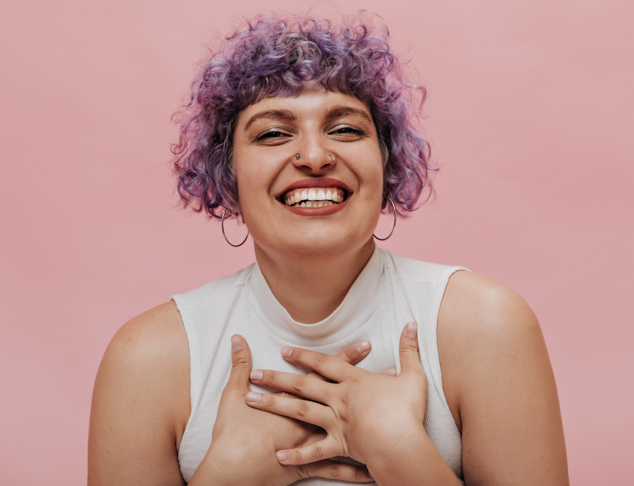 Kvinne med lilla krøllete hår, øreringer og to nesepiercinger og beige topp, smiler inn i kameraet og holder seg på brystet foran rosa bakgrunn.