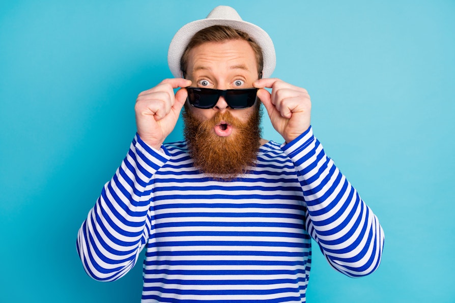Mann med rødlig skjegg blå- og hvitstripete genser og hvit hatt skyver solbrillene ned på nesen og ser overrasket ut foran lyseblå bakgrunn.