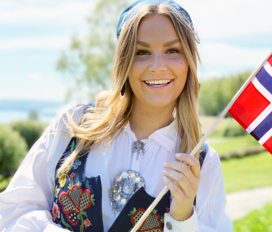 Kvinne med langt hår i bunad holder det norske flagget og smiler.