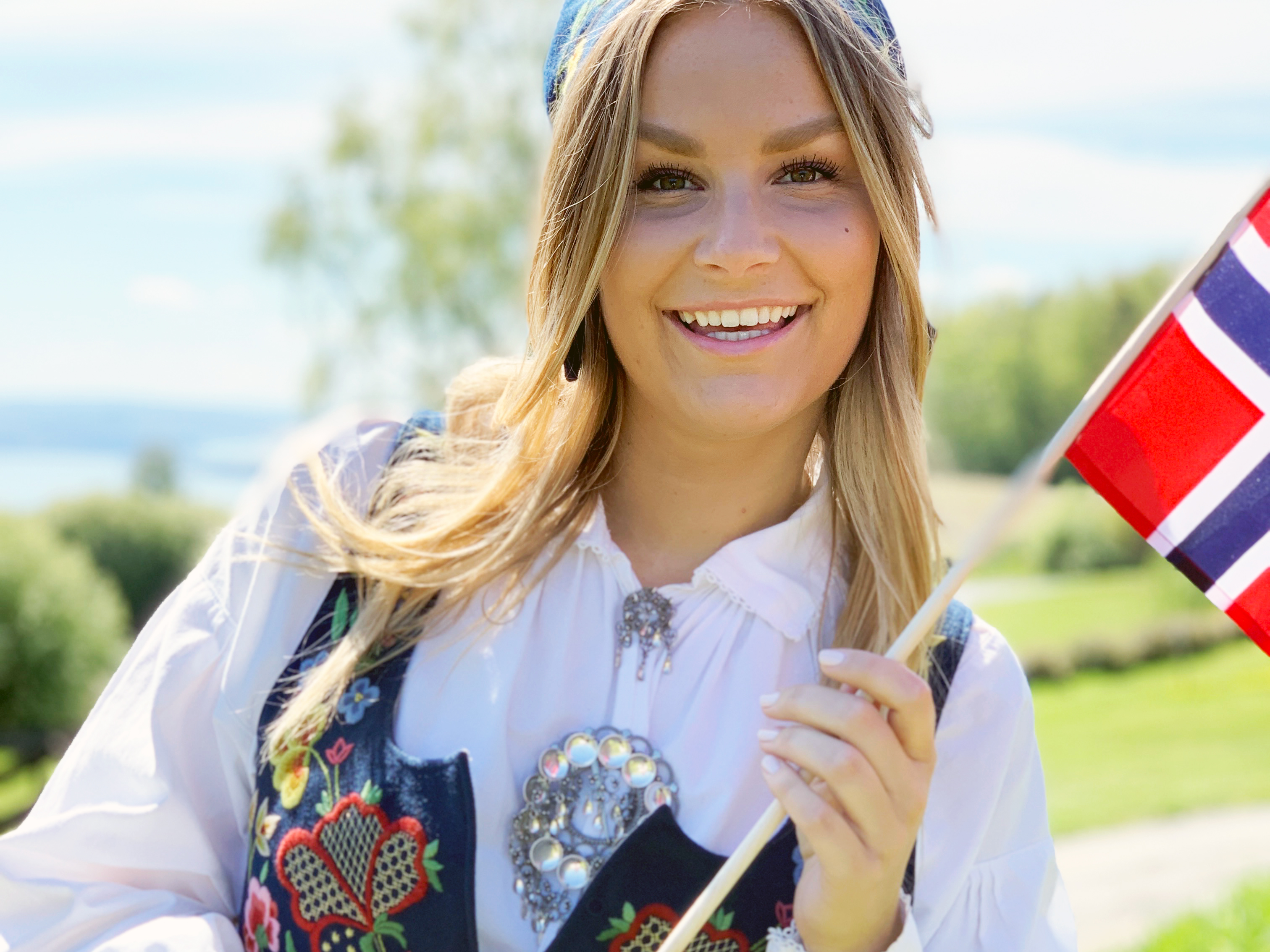 Kvinne med langt hår i bunad holder det norske flagget og smiler.