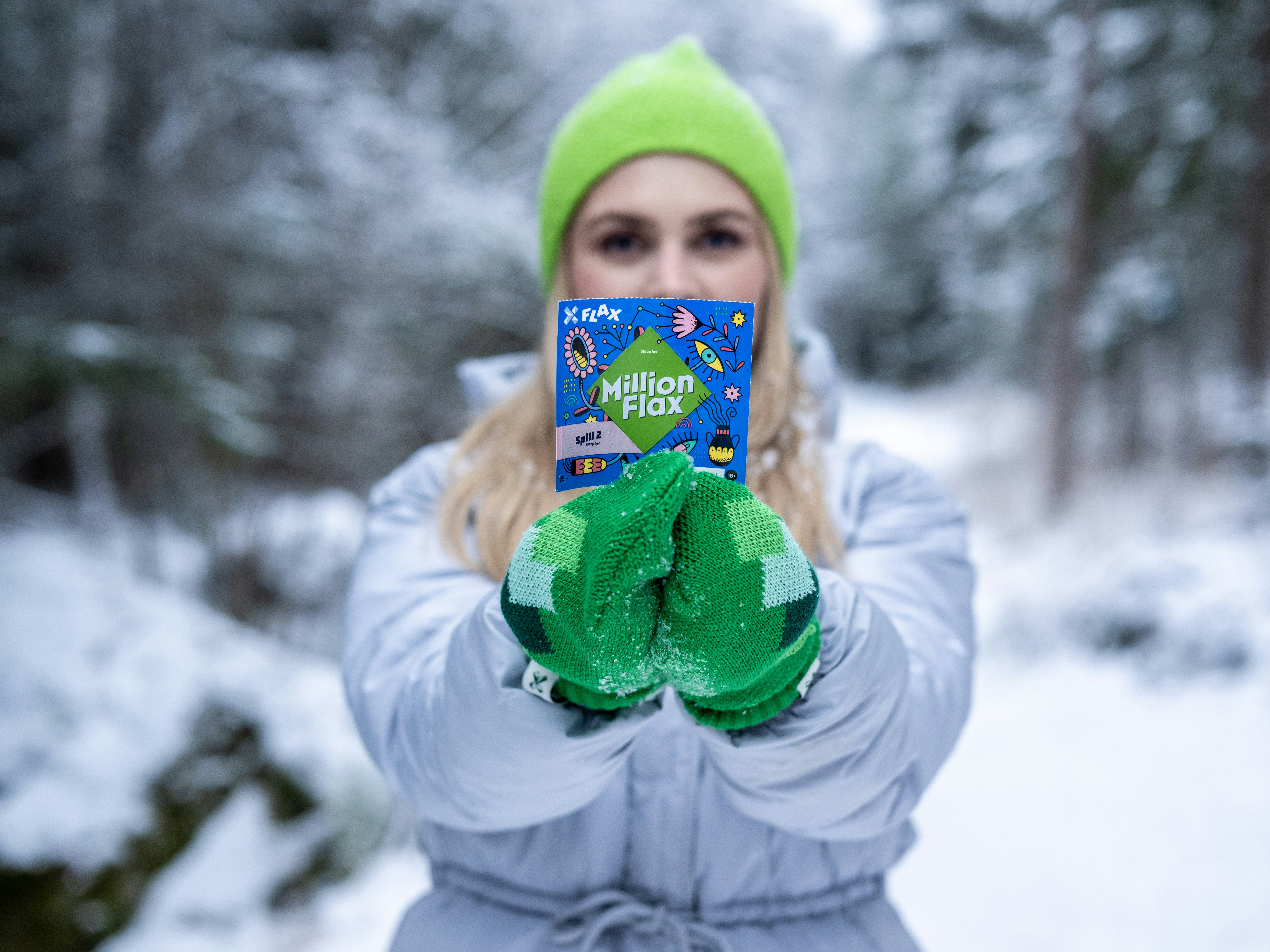 Kvinne i grønn lue, grønne votter og lyseblå boblejakke holder frem et MillionFlax-lodd med begge hendene foran vinterskog.