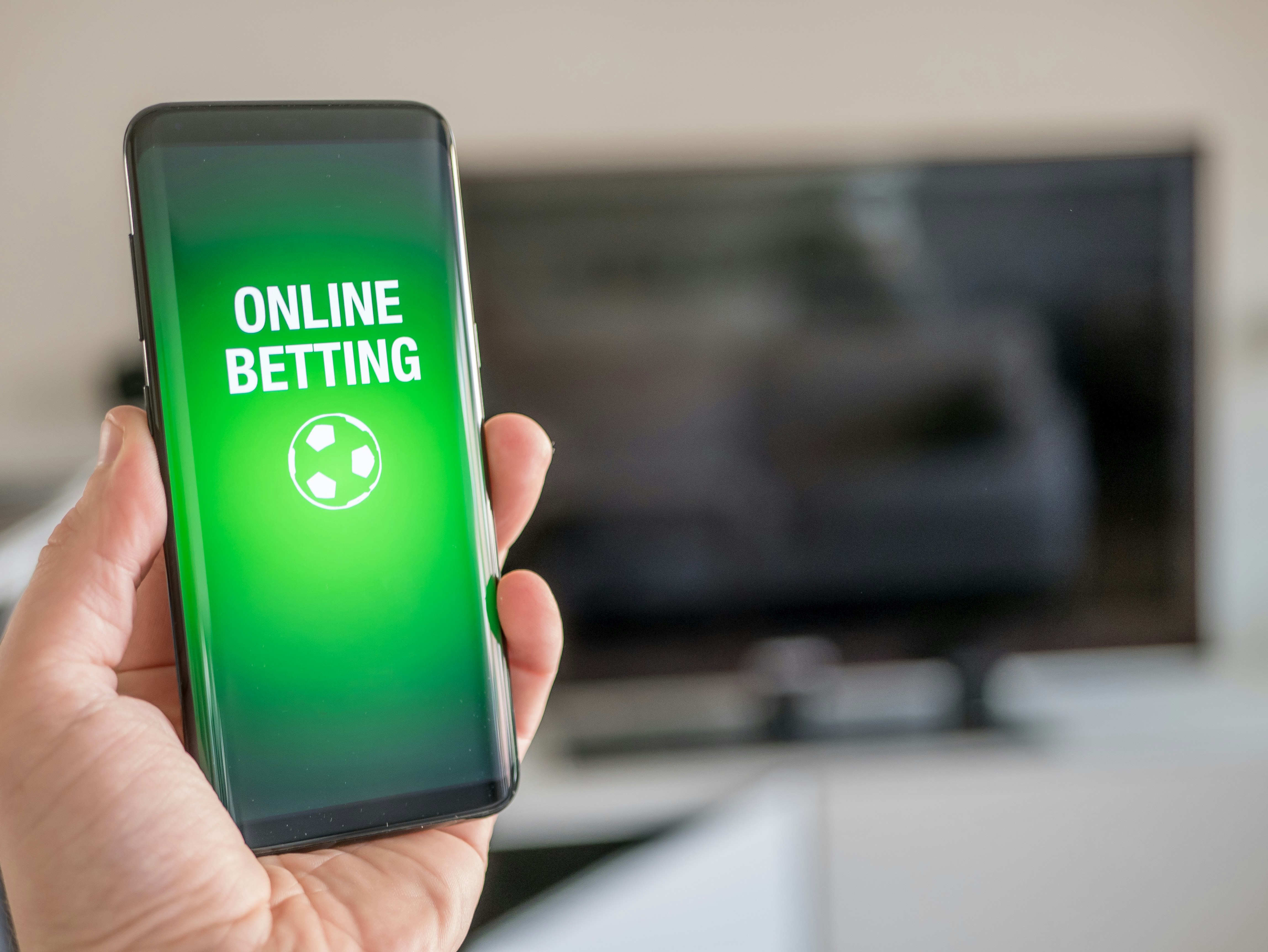 En hånd som holder en mobiltelefon der displayet er grønt, med hvit tekst som sier Online Betting, med tegning av en fotball under.